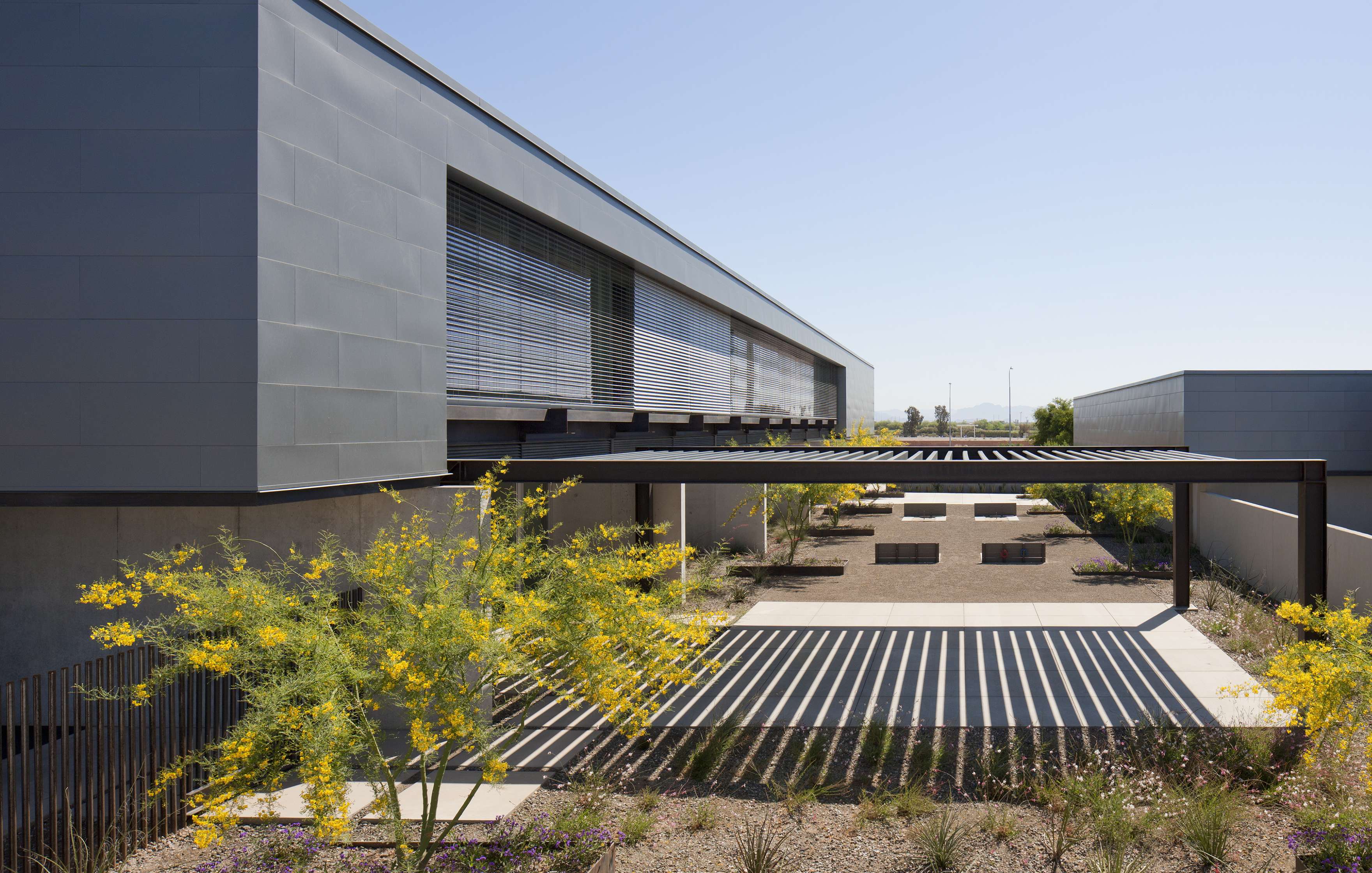 Modern building with large horizontal windows and metal slatted awnings casting shadows over a landscaped courtyard with trees and pathways.