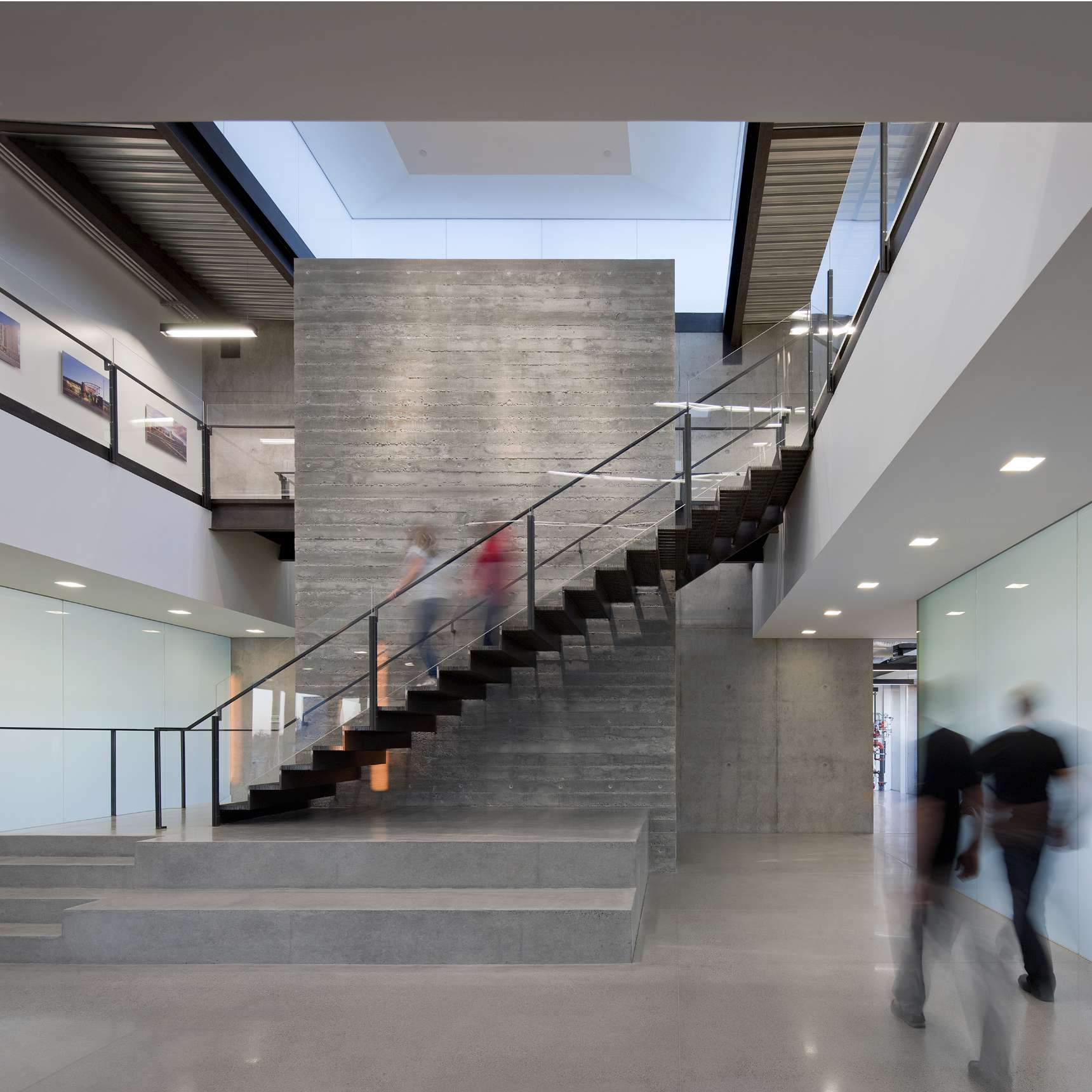 Modern interior with a concrete staircase and glass balustrades. People are walking up and down the stairs and through the corridor. Sleek, minimalist design with large ceiling light.