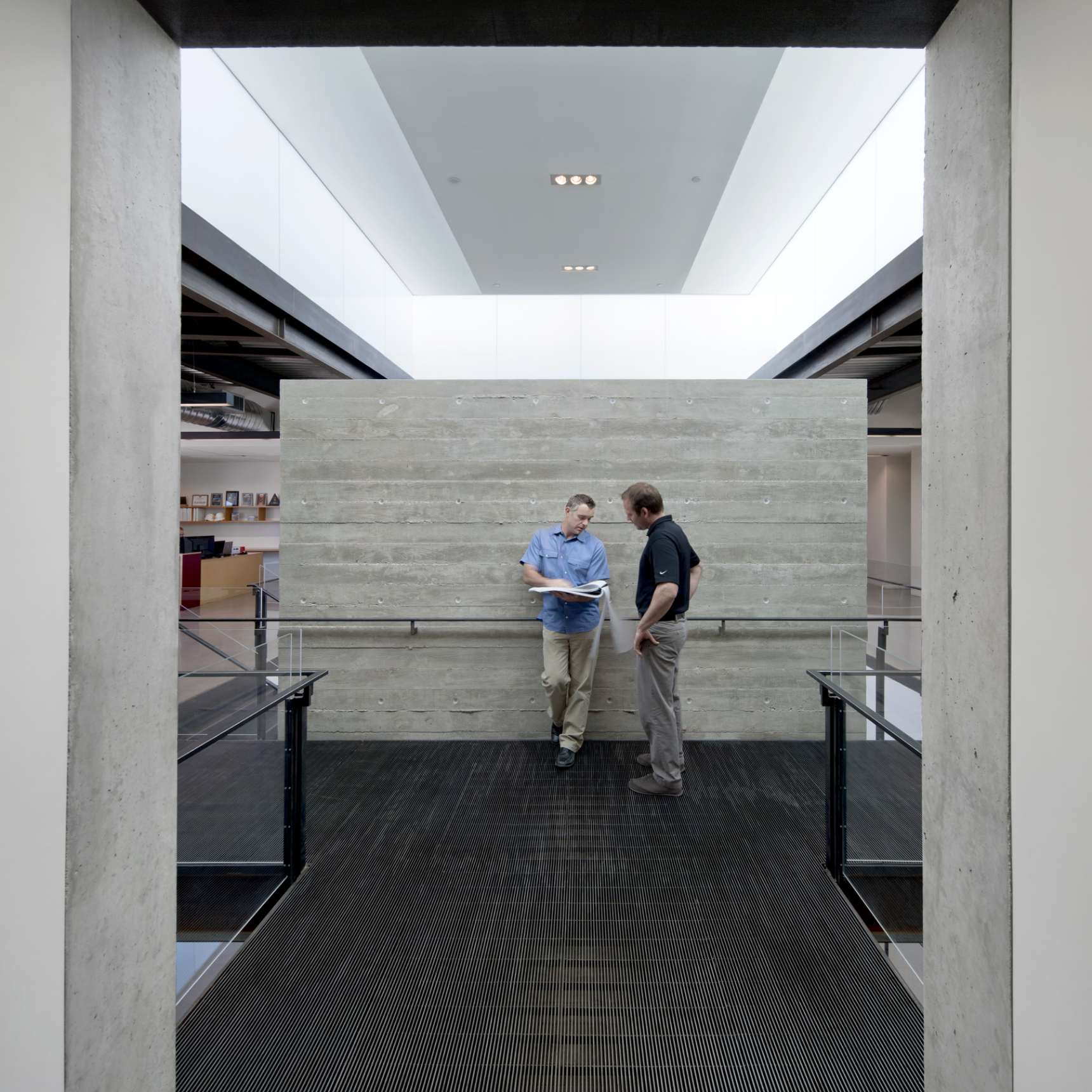 Two men stand talking on a walkway inside a modern building with a concrete wall and open two-story design.