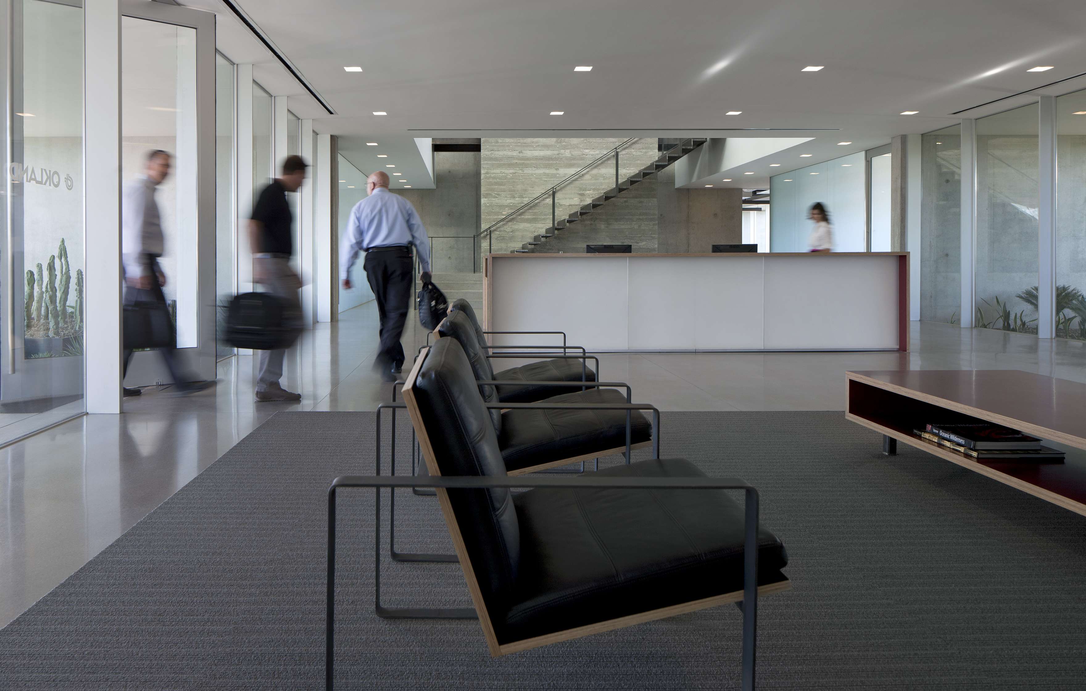 A modern office lobby with black chairs, a low table, and people walking briskly; a staircase and reception desk are visible in the background.