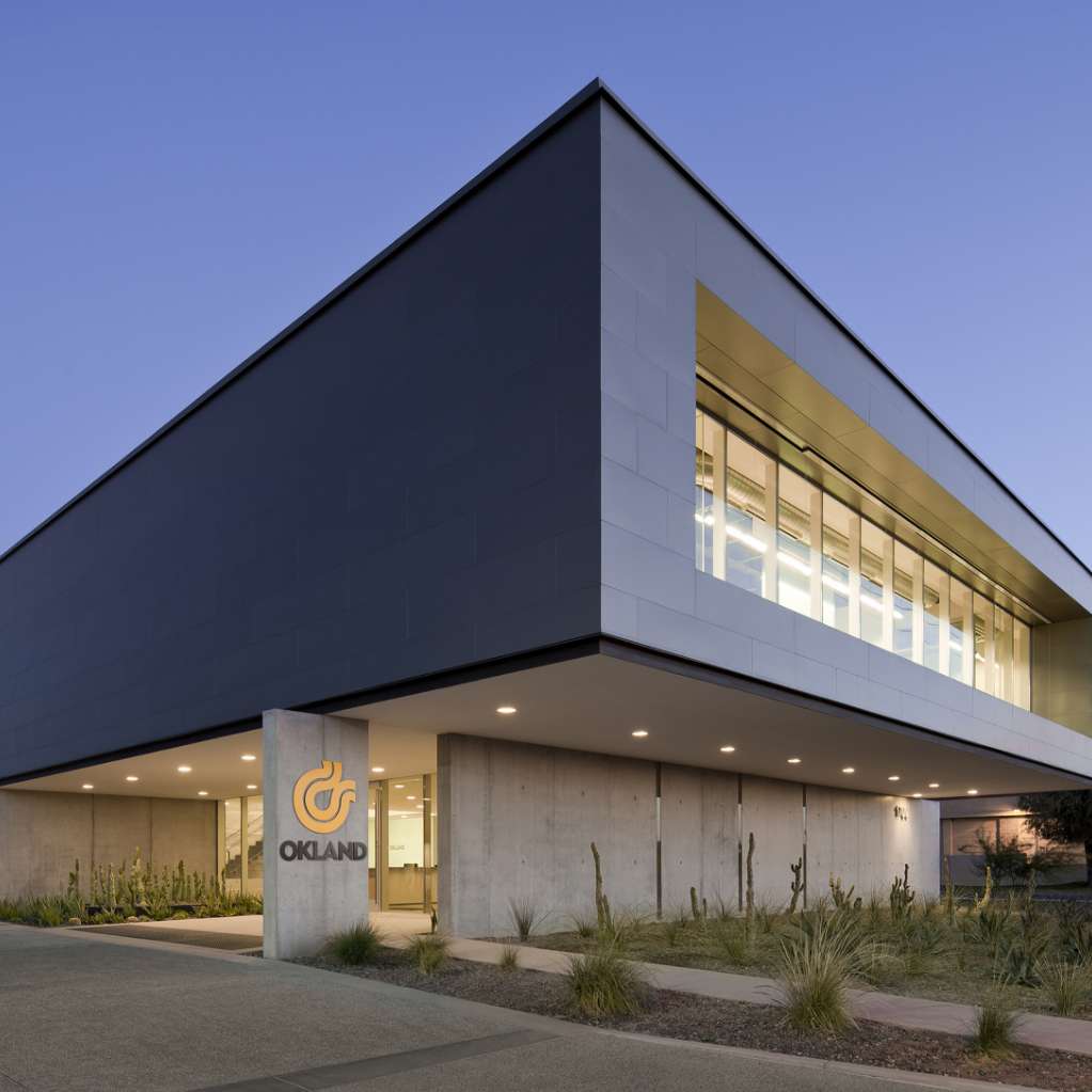 Modern building with large windows and a sign that reads “OKLAND” near the entrance, surrounded by desert landscaping. The sky is clear, suggesting it is either early morning or late evening.