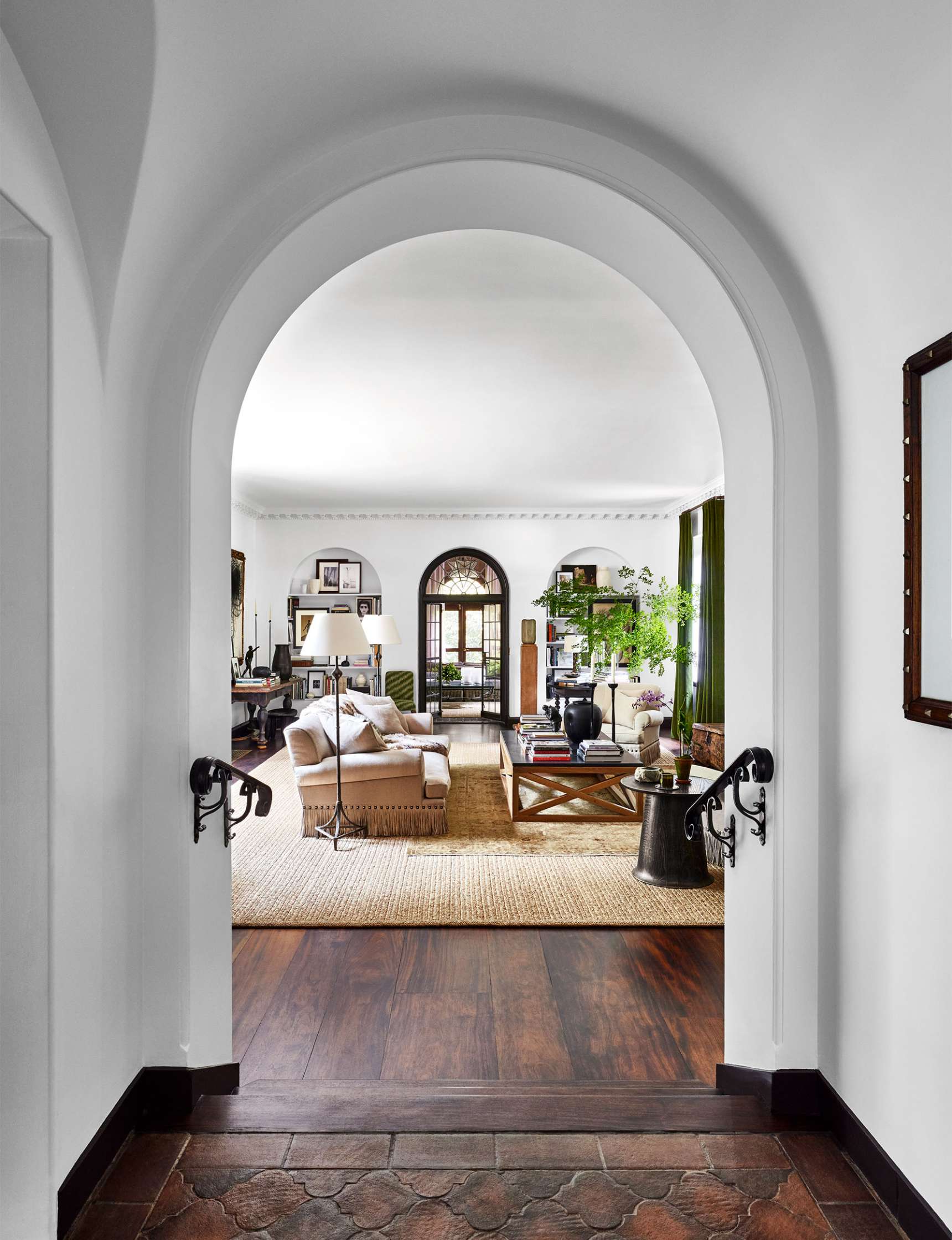 View through an arched doorway into a living room with a beige sofa, ceiling lamp, coffee table, rug, and greenery. Large windows let in natural light. Dark wood floor contrasts with white walls.