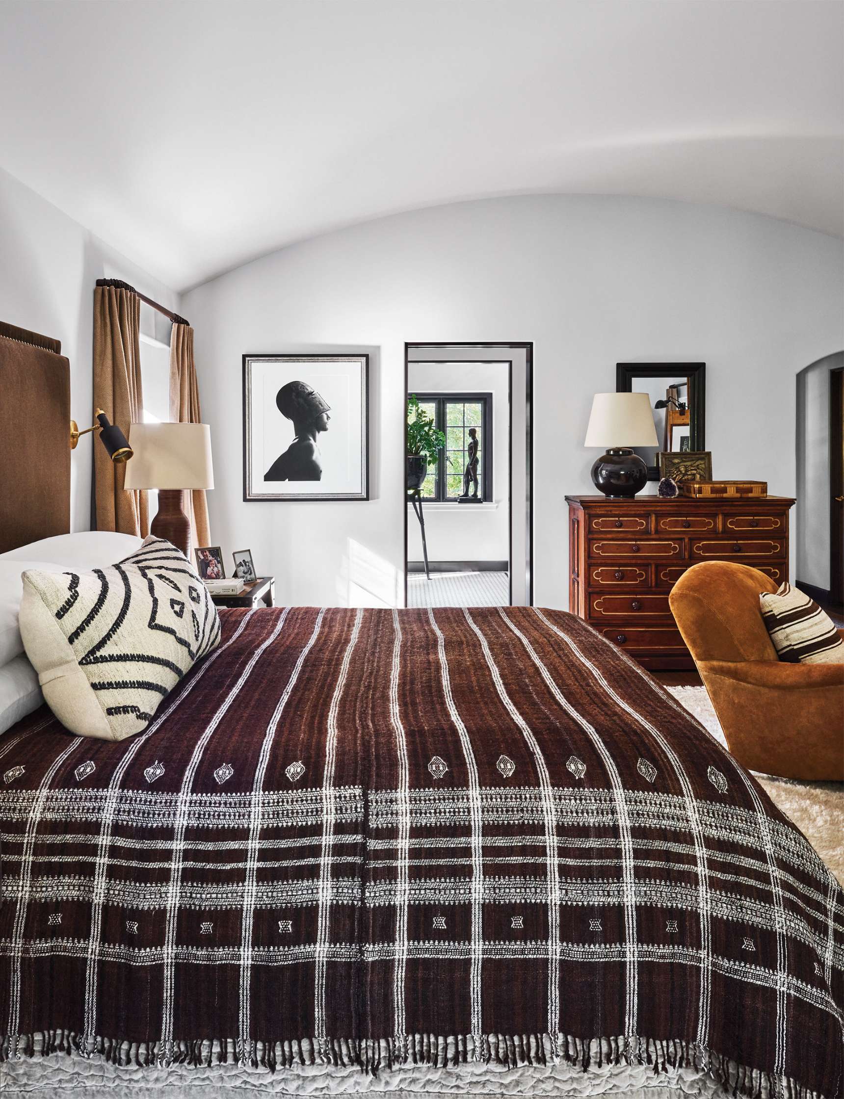 A bedroom with a brown patterned bedspread, a wooden dresser, an armchair, and framed artwork on the wall. A large mirror reflects a window with greenery outside.