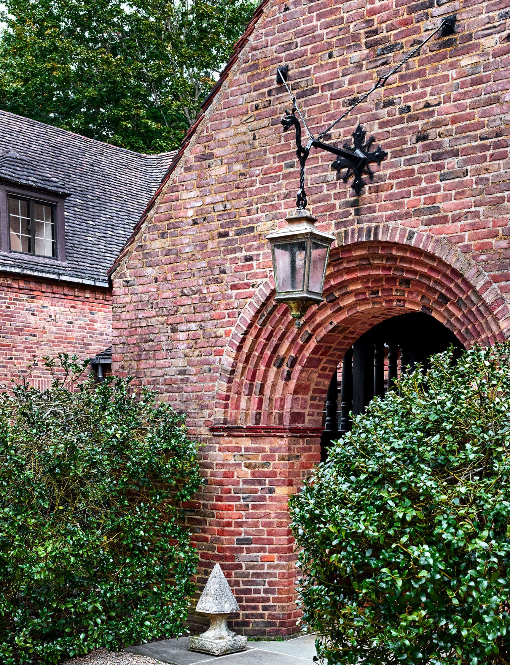 Brick building with arched entrance, lantern hanging above, surrounded by green shrubs and trees.