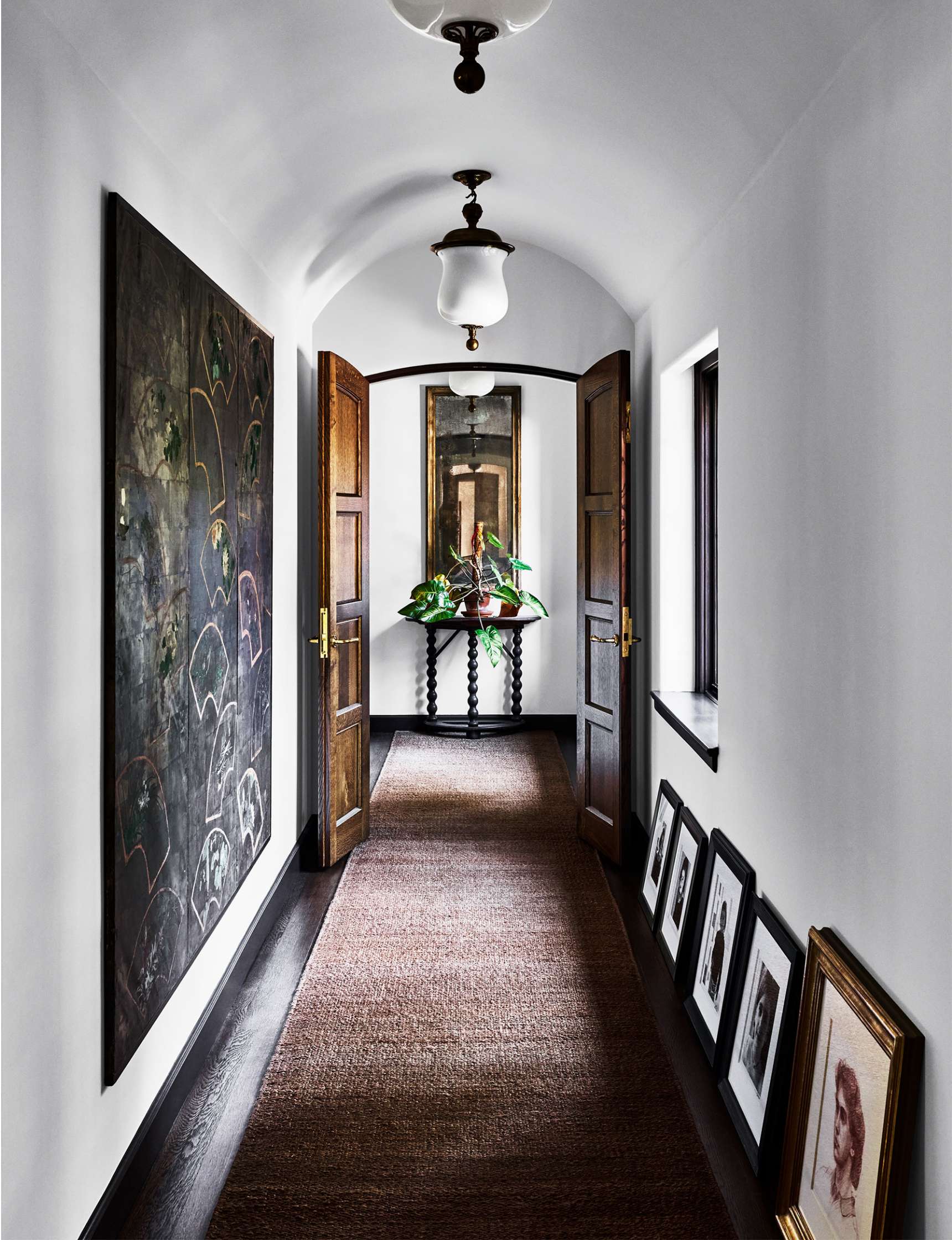 A narrow hallway with framed art on the walls and floor leading to a small table with a decorative plant and a mirror above. The floor is covered with a natural fiber rug.