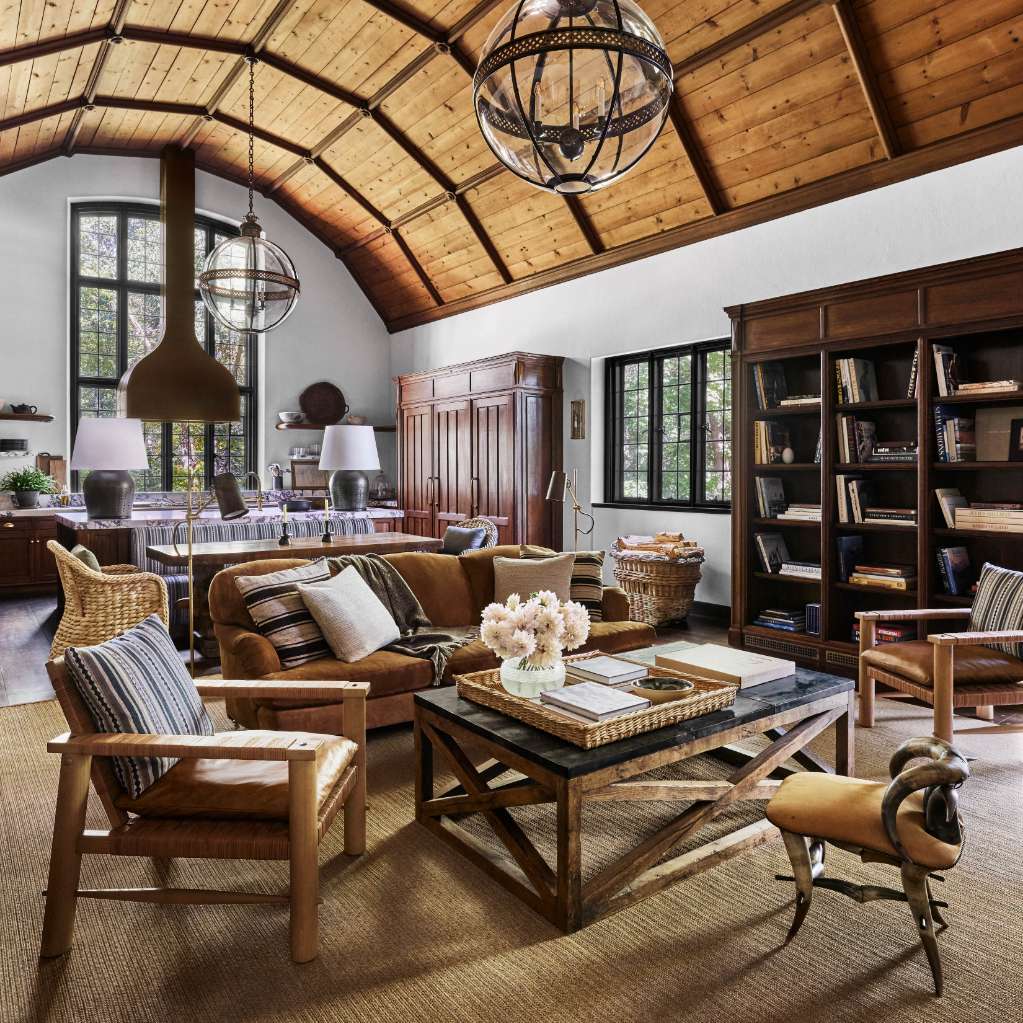 A rustic living room with a high wooden ceiling, leather sofas, a wooden coffee table, bookshelves, large windows, and two spherical chandeliers.