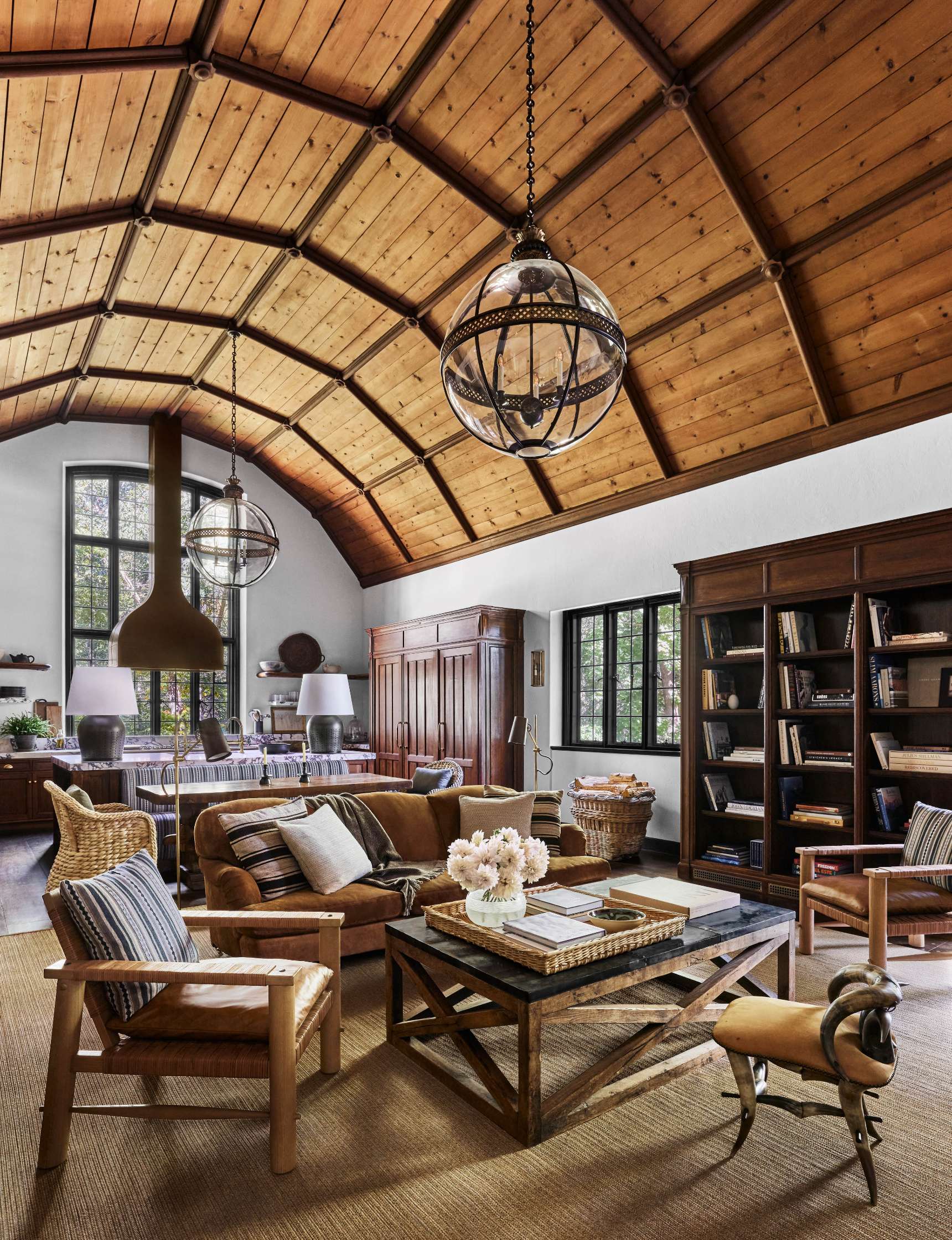 A rustic living room with a high wooden ceiling, leather sofas, a wooden coffee table, bookshelves, large windows, and two spherical chandeliers.