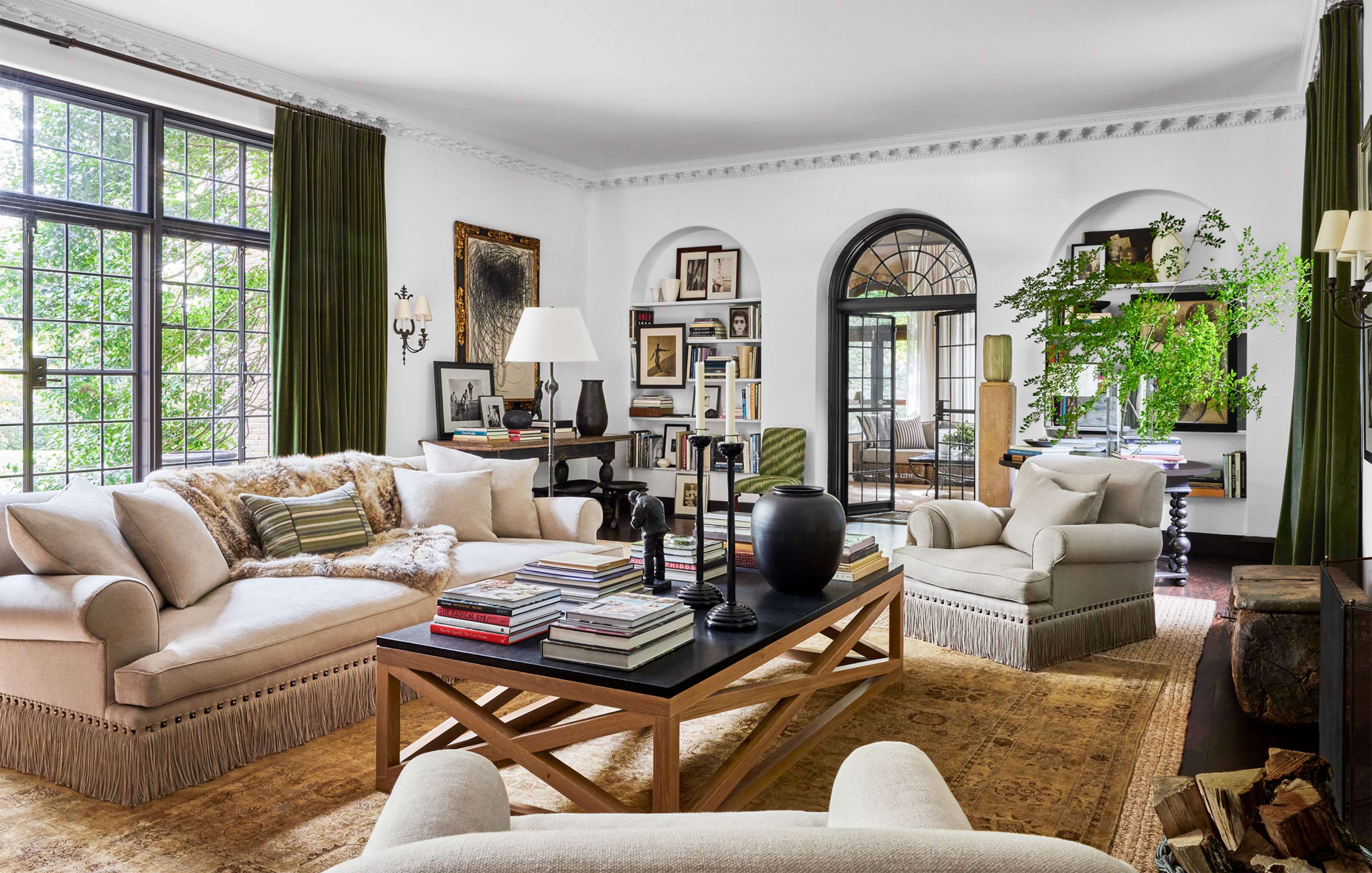 A spacious living room with cream sofas, a wooden coffee table, and books. Large windows with green curtains, decorative plants, and art pieces adorn the space.