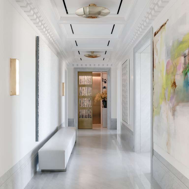 A modern, white hallway with a sleek bench on the left, abstract artwork on the walls, and recessed lighting overhead leads to a room with glass doors and decorative plants.