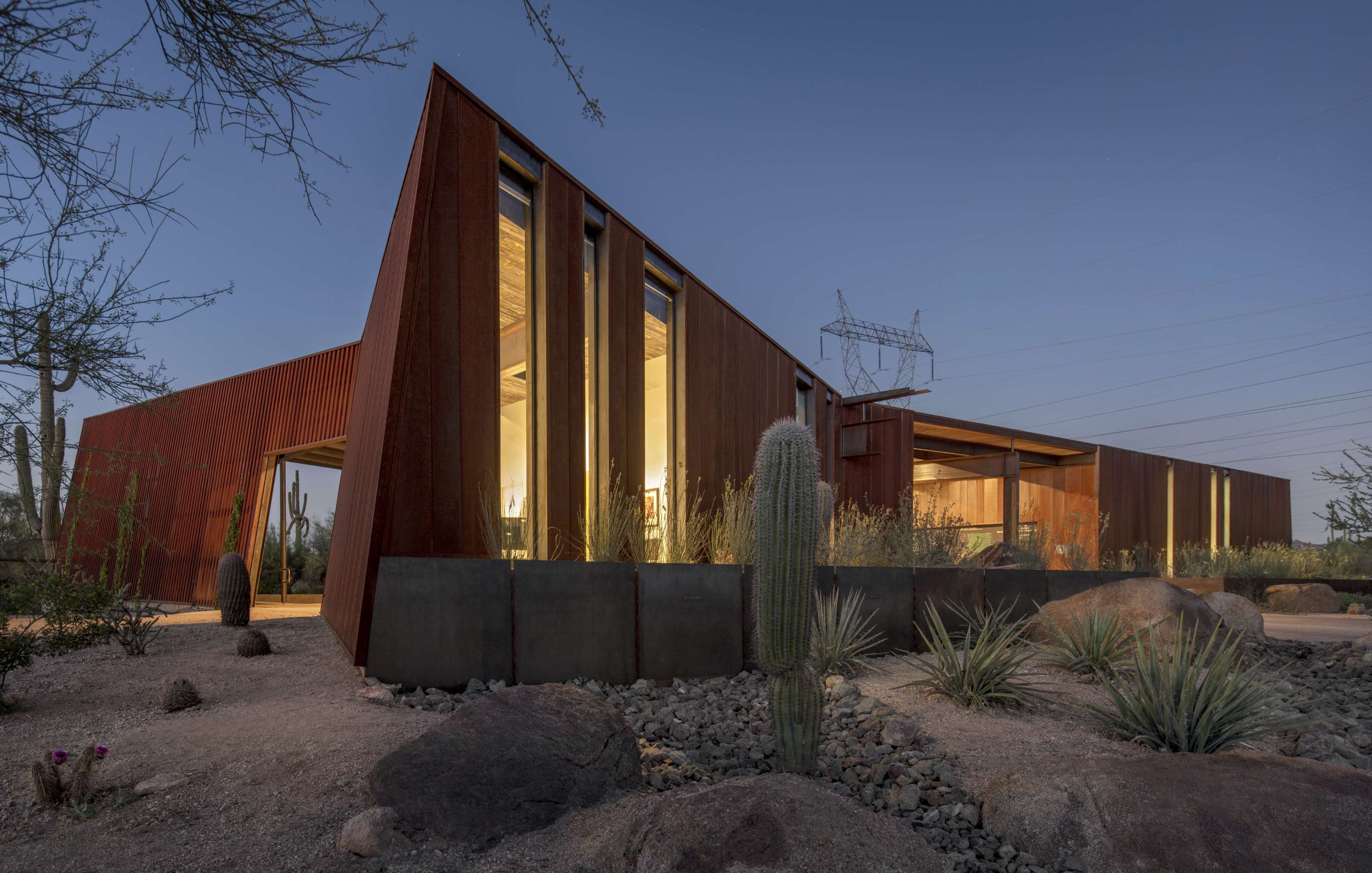 A modern, angular building with rust-colored walls and large windows, surrounded by desert plants and cacti, under a twilight sky.