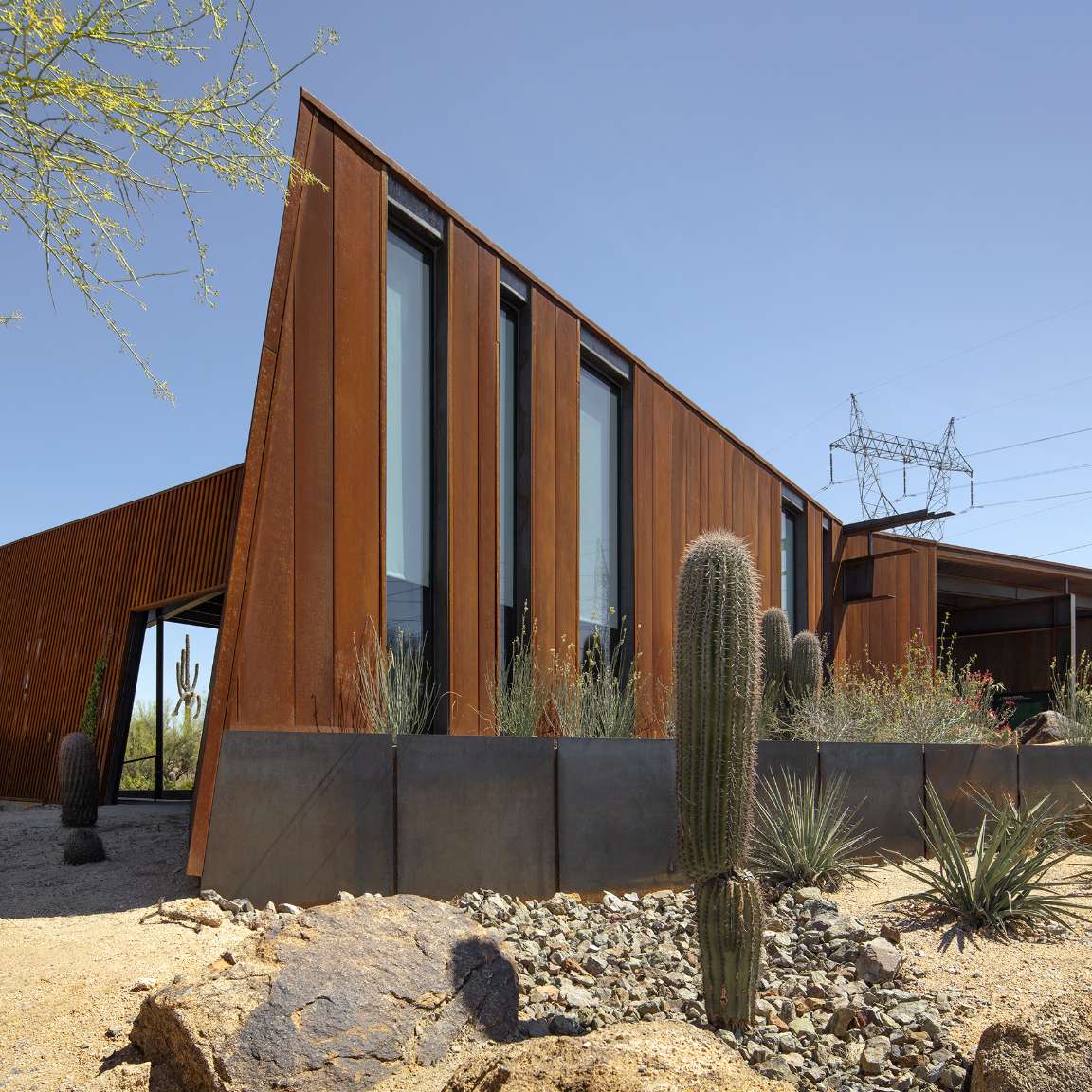 Modern building with rust-colored metal panels and large windows in a desert landscape, surrounded by cacti and sparse vegetation. Power lines are visible in the background.