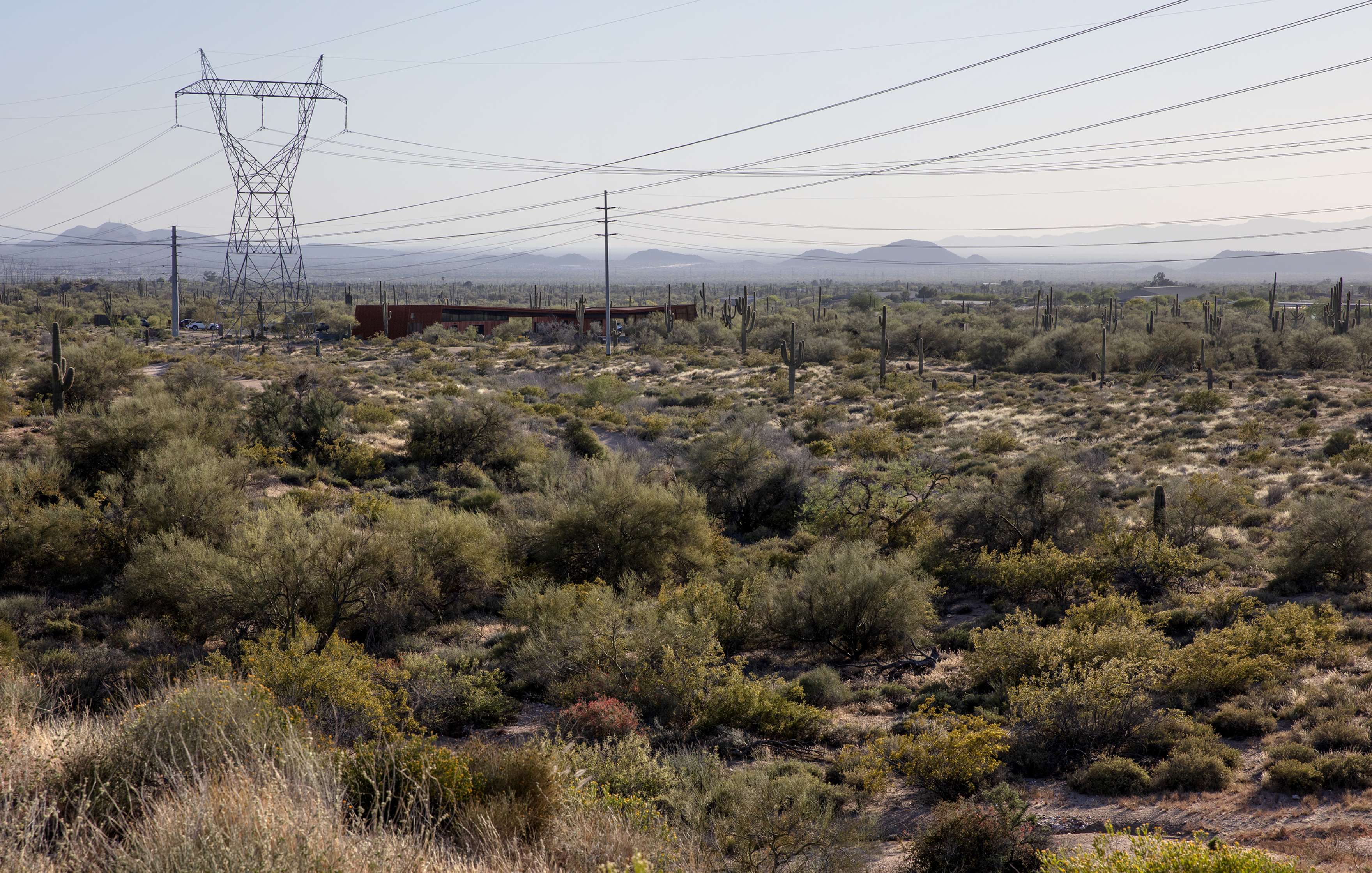 A desert landscape with sparse vegetation, power lines, and a large transmission tower. The background features distant mountain ranges under a clear sky.