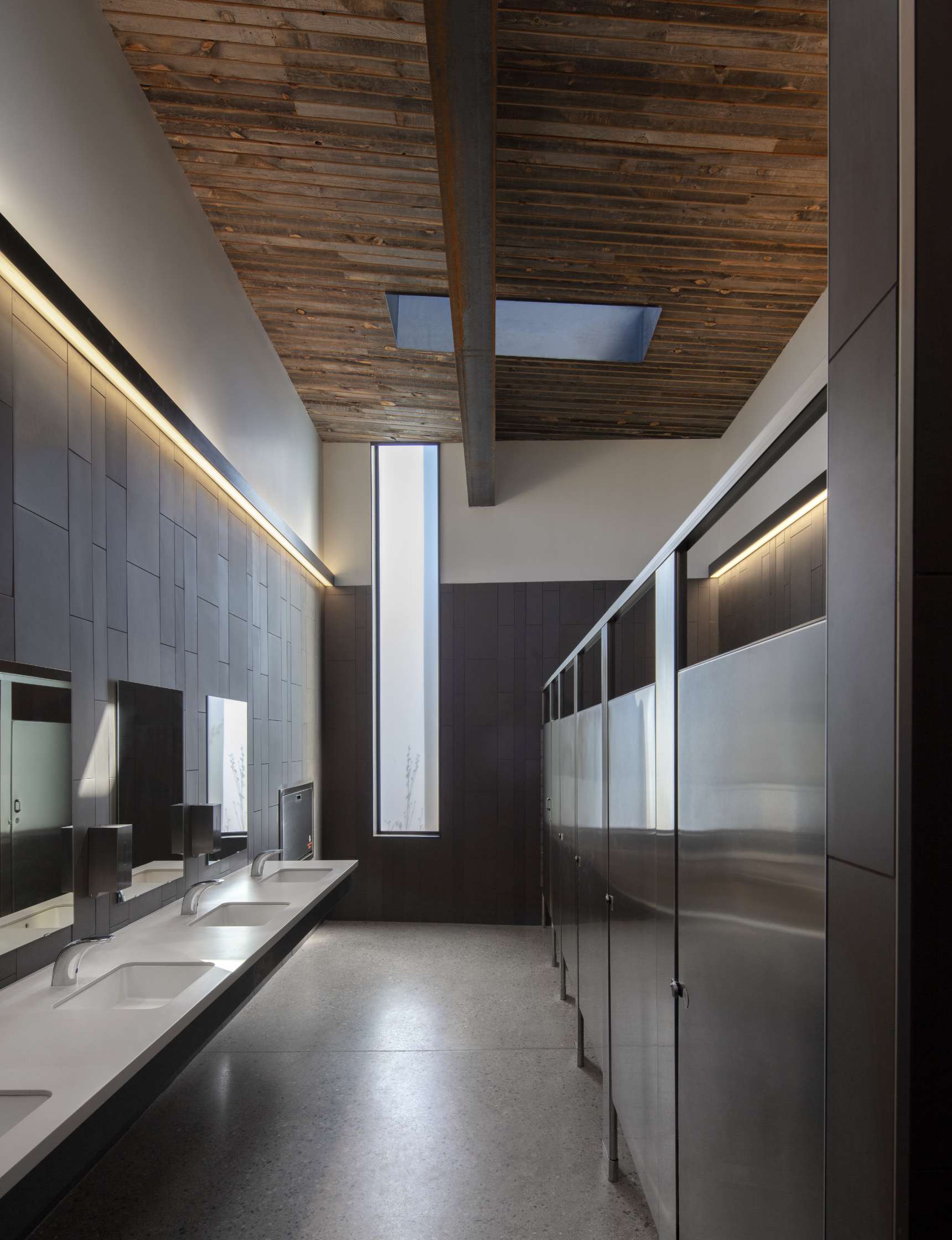 A modern restroom with a wooden ceiling, skylight, and tall windows. It features a long countertop with sinks and mirrors on the left and metal stalls on the right.