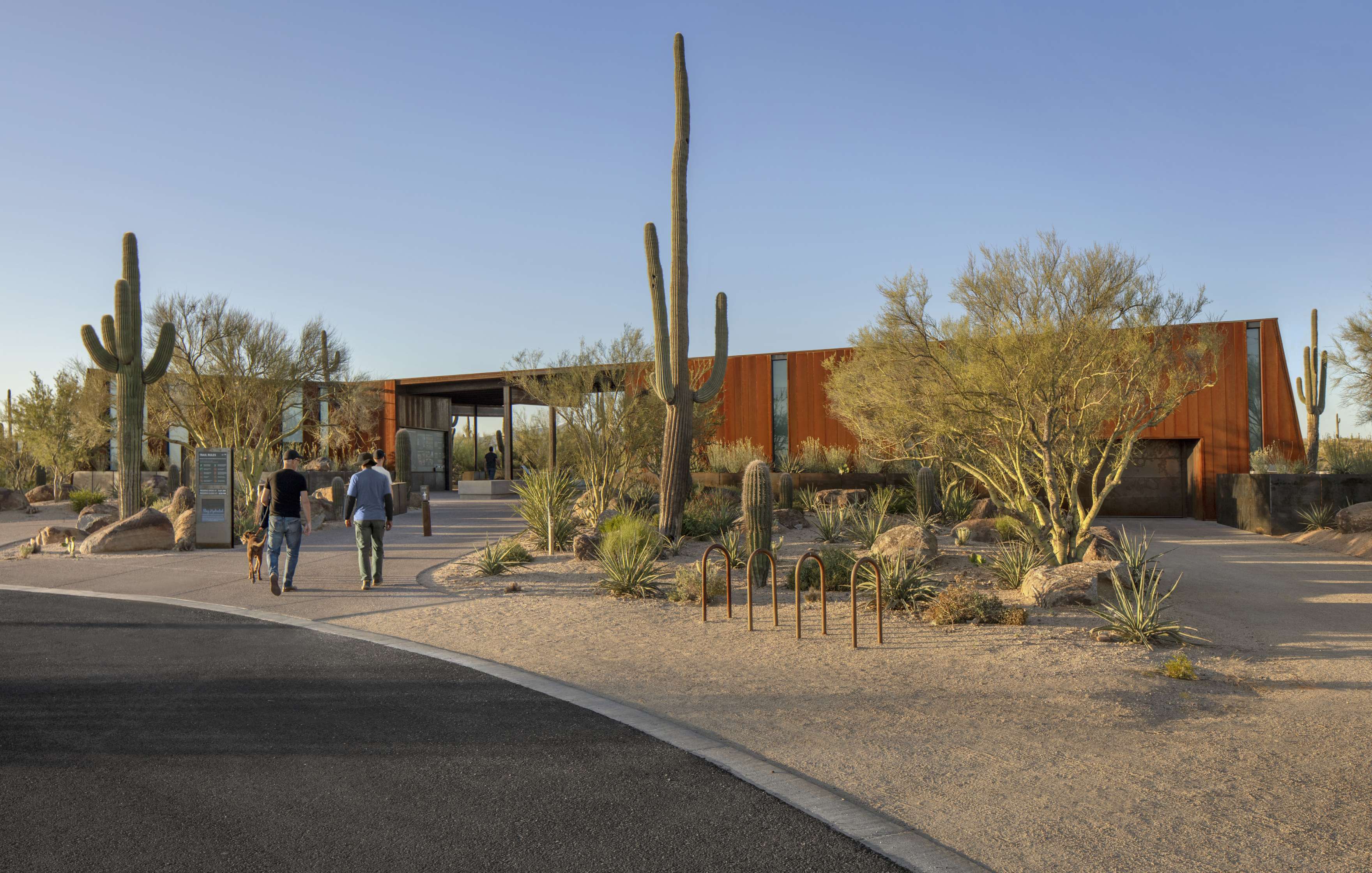 Three people walk towards a modern building surrounded by cacti and desert vegetation, under a clear sky.