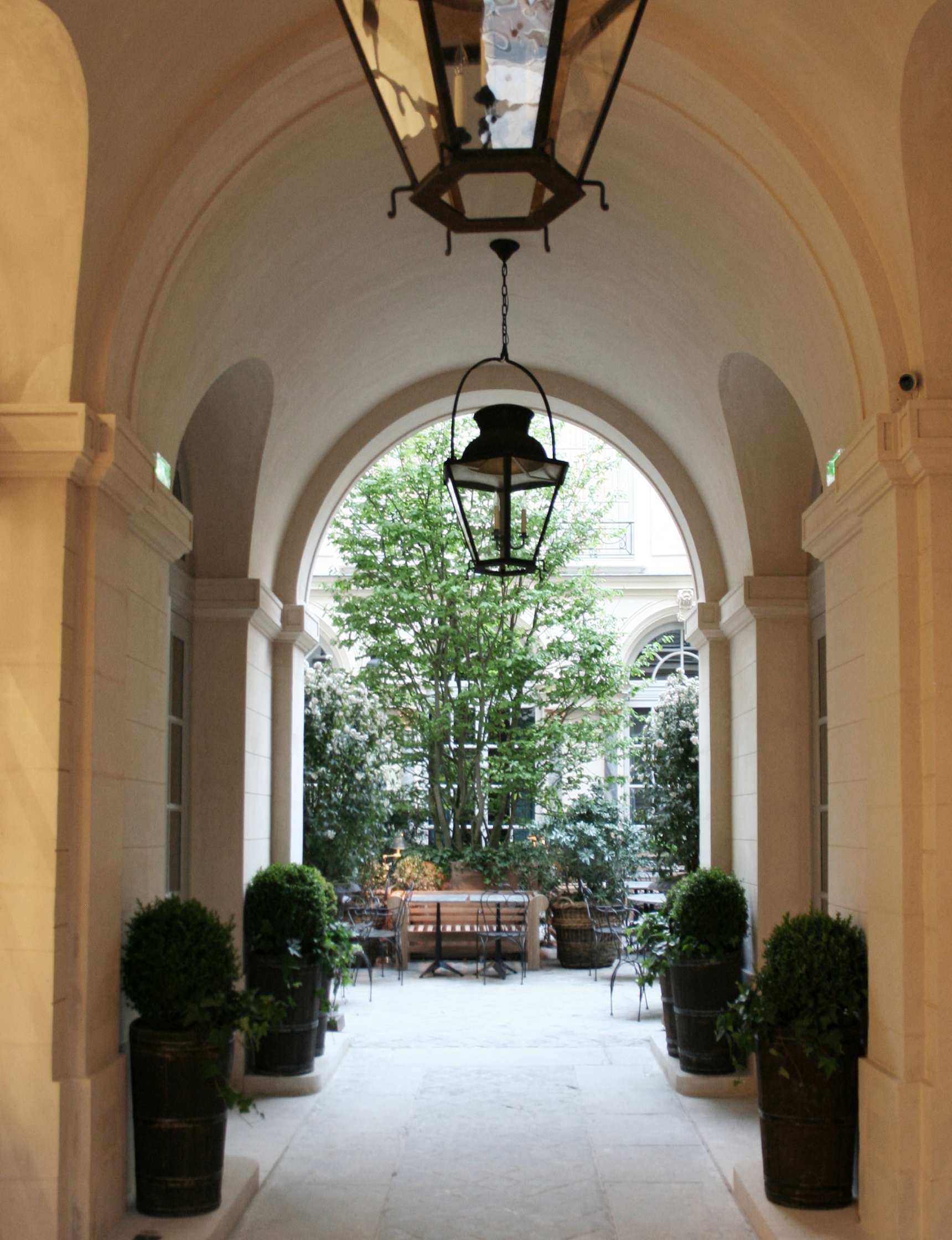 A covered archway leads to a small courtyard with a wooden bench, surrounded by potted plants and greenery, under soft light from an overhead lantern.