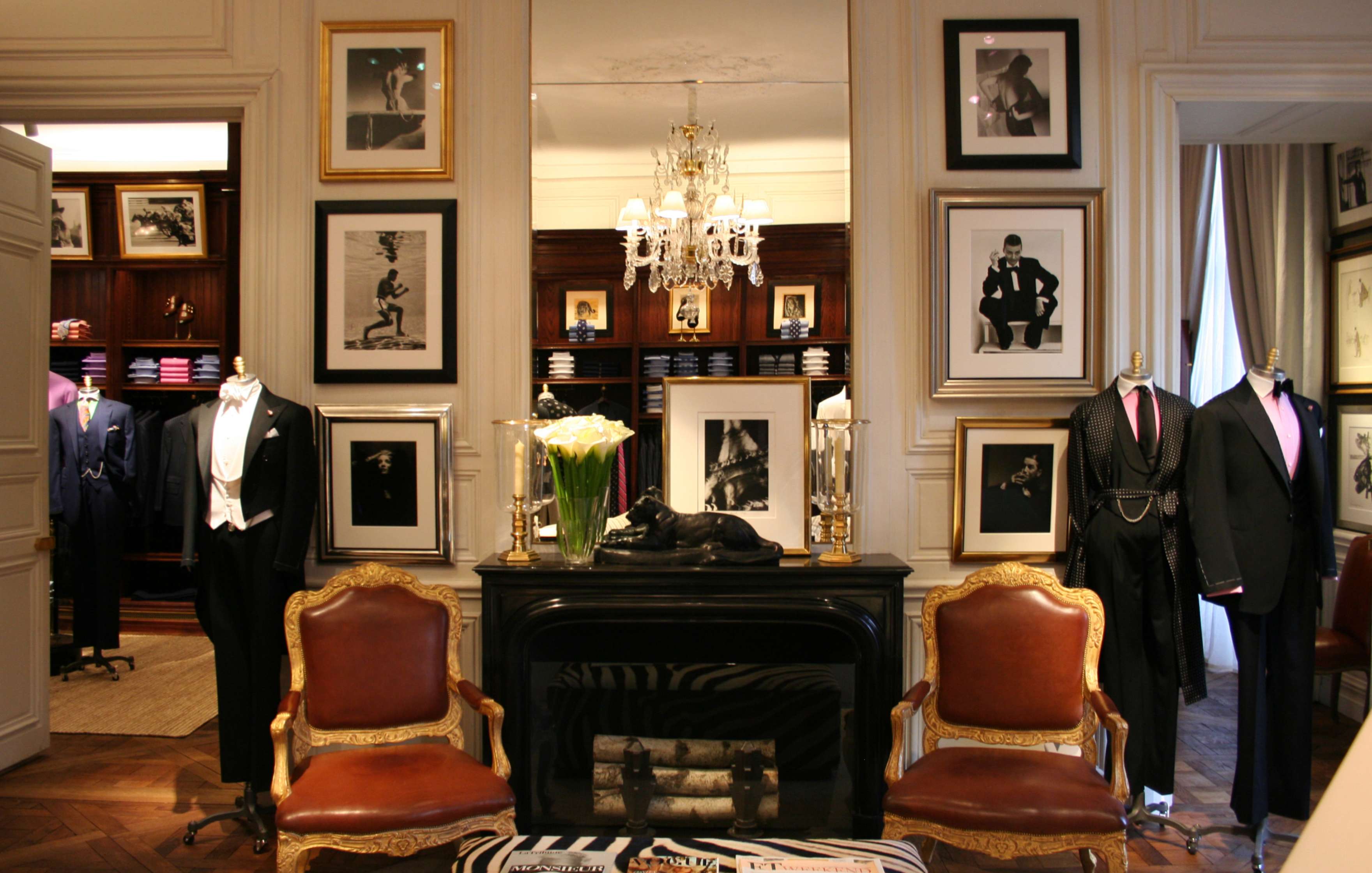 A well-lit, elegant men’s clothing store showcases tuxedos and suits on mannequins, with framed photos and a chandelier enhancing the sophisticated decor.