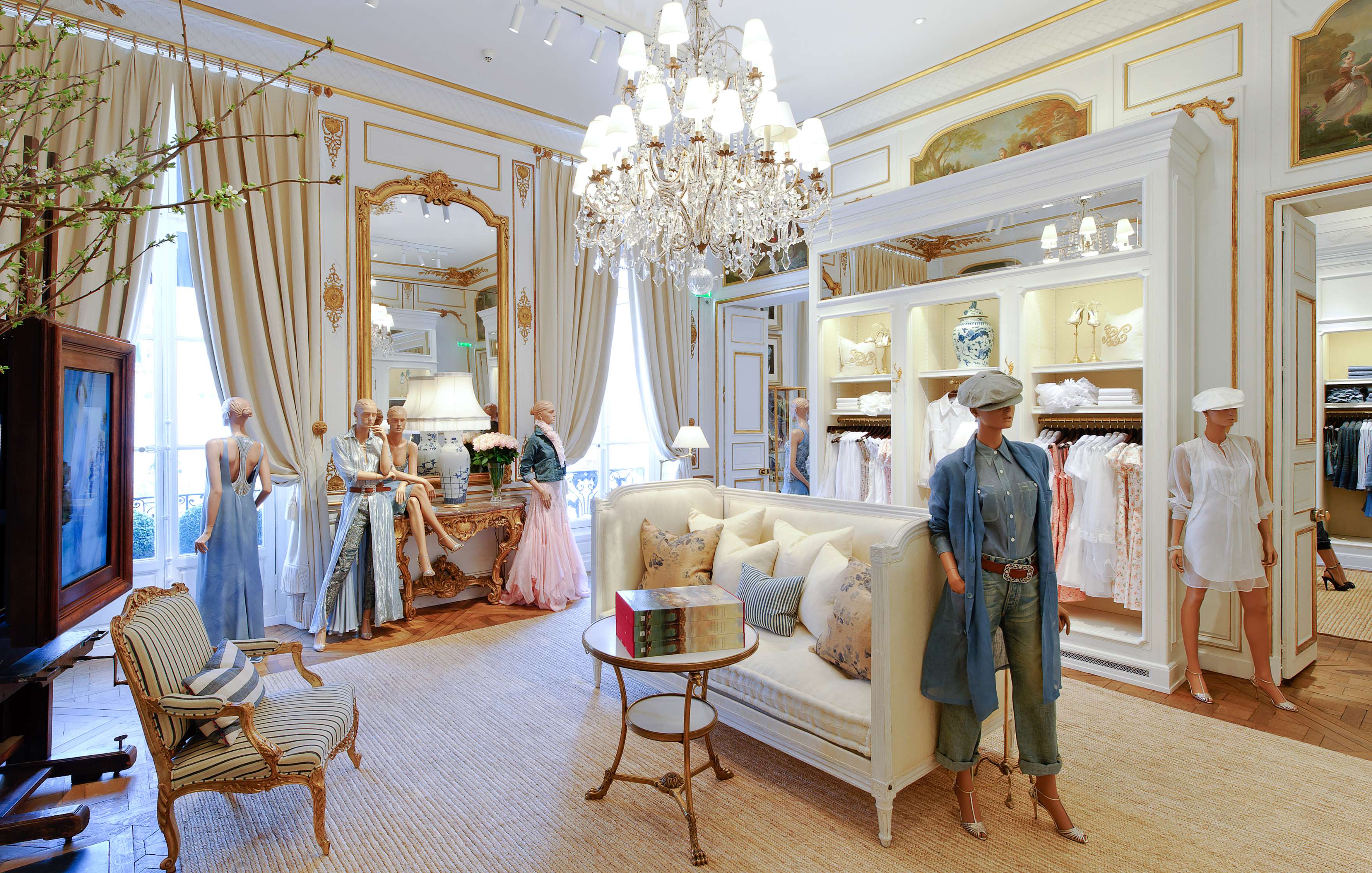 Luxurious boutique interior with elegant decor, showcasing mannequins in fashionable clothing, a chandelier, mirrors, and a central seating area with a table. Clothing items are neatly displayed on shelves and racks.