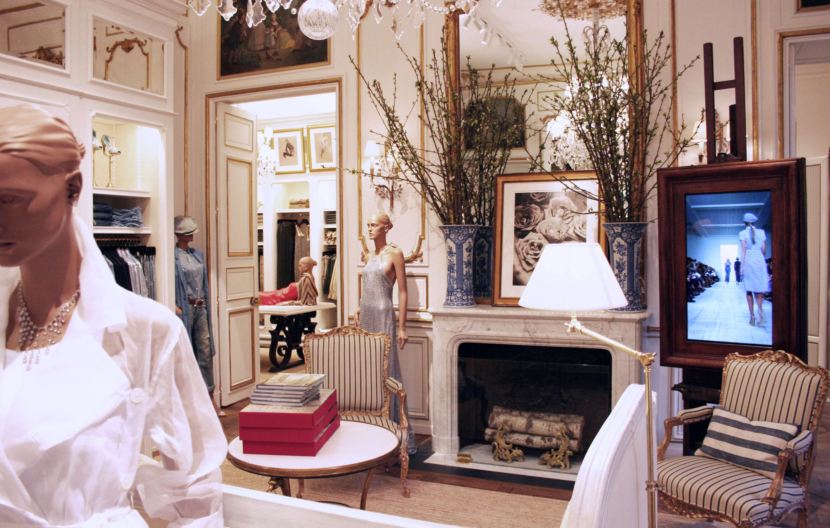 A luxurious clothing store interior featuring mannequins in white attire, ornate decor, a fireplace with elegant vases, a chandelier, and framed artwork on the walls.