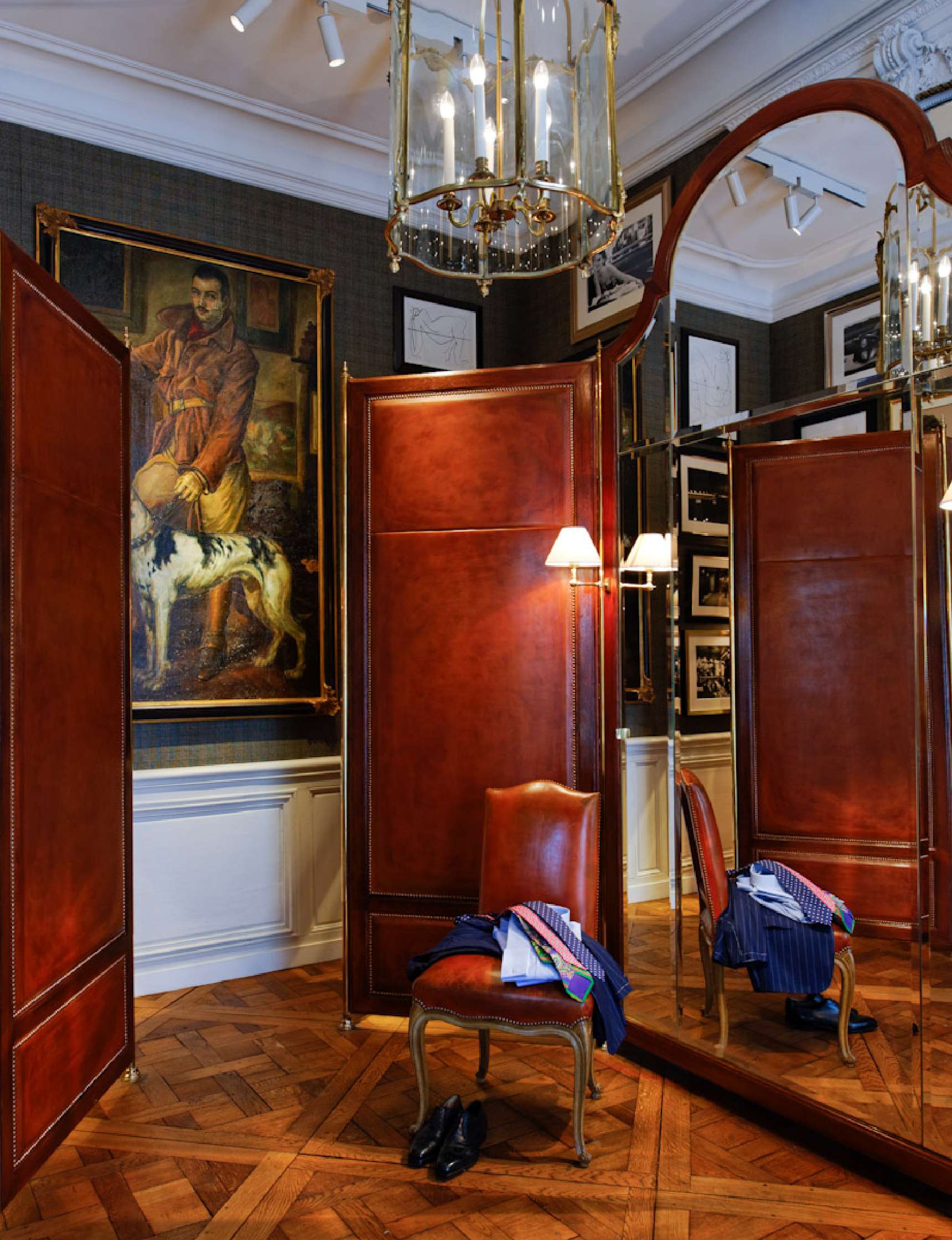 Luxurious dressing room with leather-paneled walls, large mirrors, a chandelier, an upholstered chair, scattered clothes, and a large painting of a man with a dog.