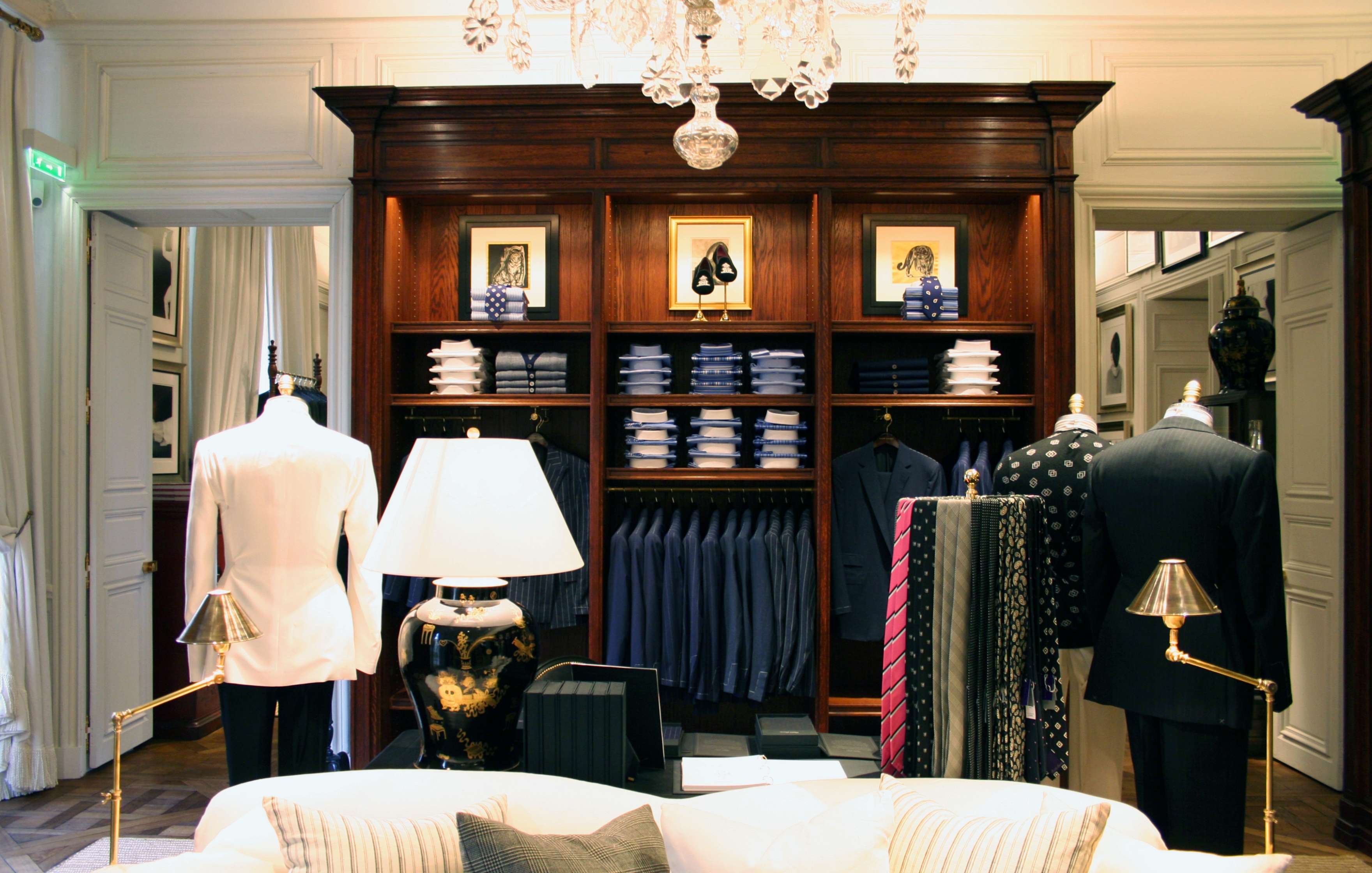 A luxury clothing store interior featuring tailored suits, ties, and folded shirts displayed on wooden shelves, with suit-clad mannequins, a black lamp, and brass lighting accents.