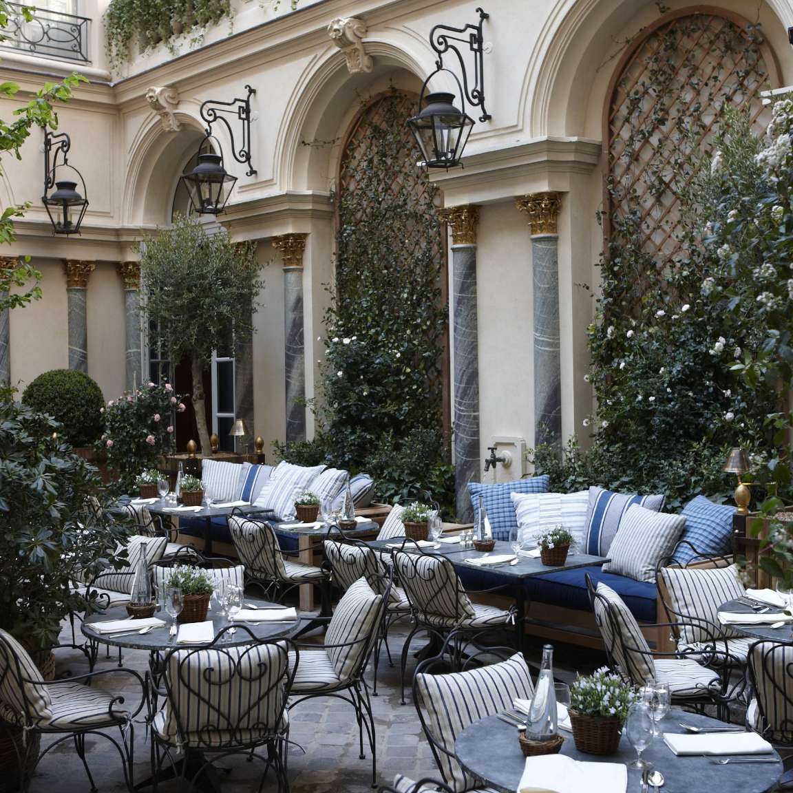 Outdoor patio set with elegant wrought-iron tables and chairs, cushioned seating, potted plants, and lush greenery against an ornate architectural backdrop with arched doorways and hanging lanterns.