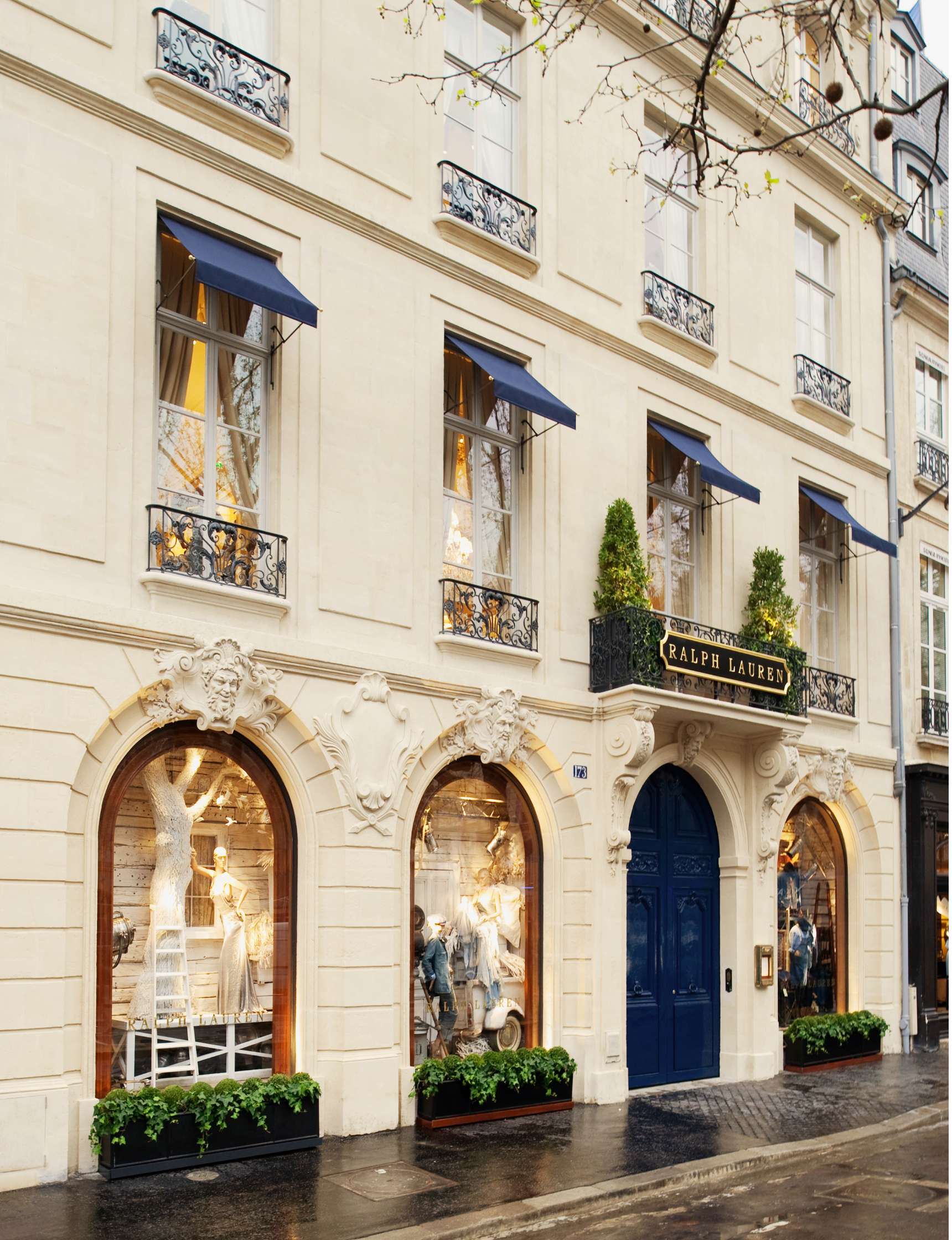 Exterior of a Ralph Lauren store with large windows displaying clothing, blue awnings, and ornate architectural details on a rainy day.