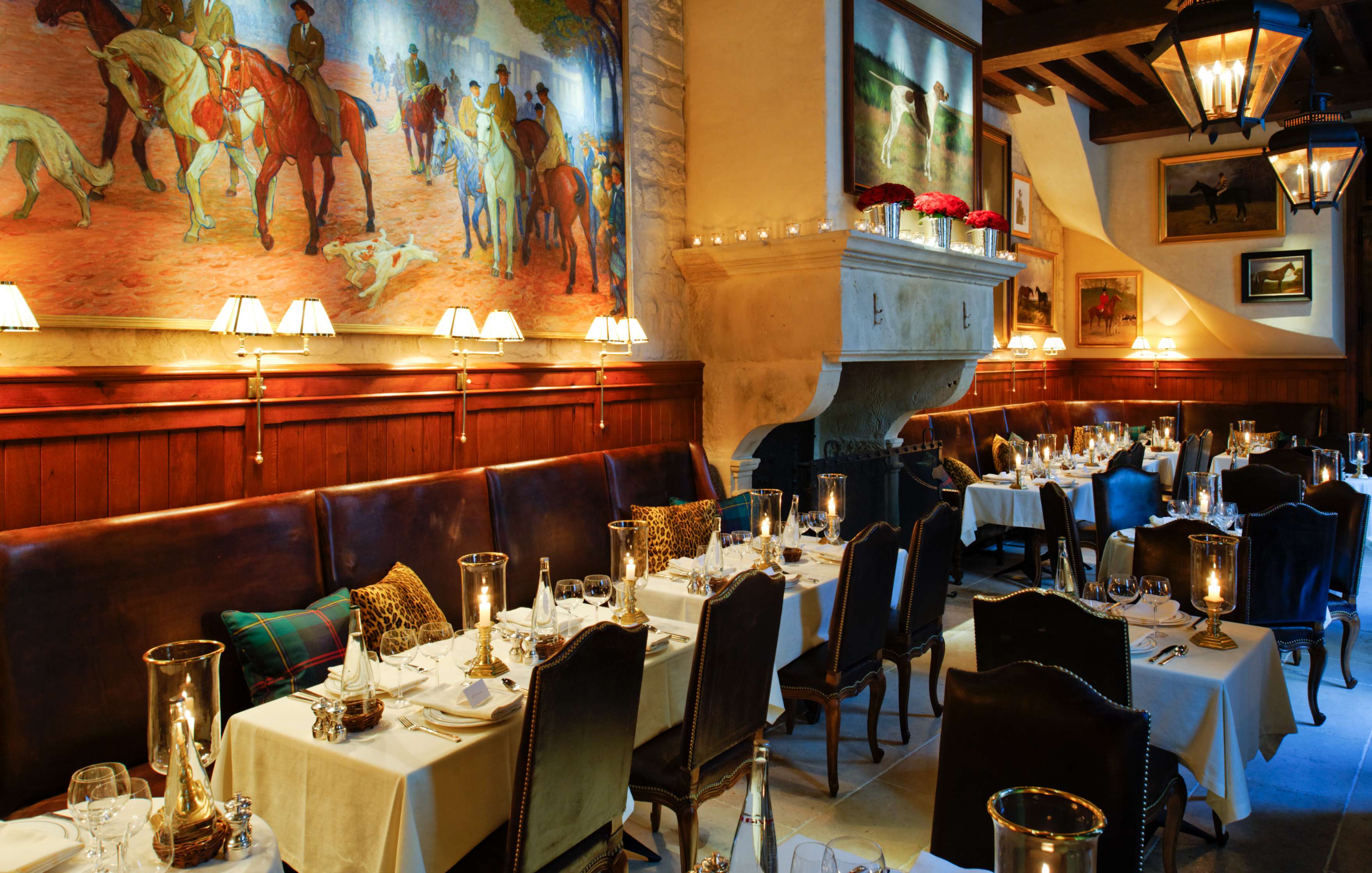 A warmly lit restaurant interior featuring white tablecloths, leather chairs, large murals of historical scenes on the walls, and a stone fireplace with candles.