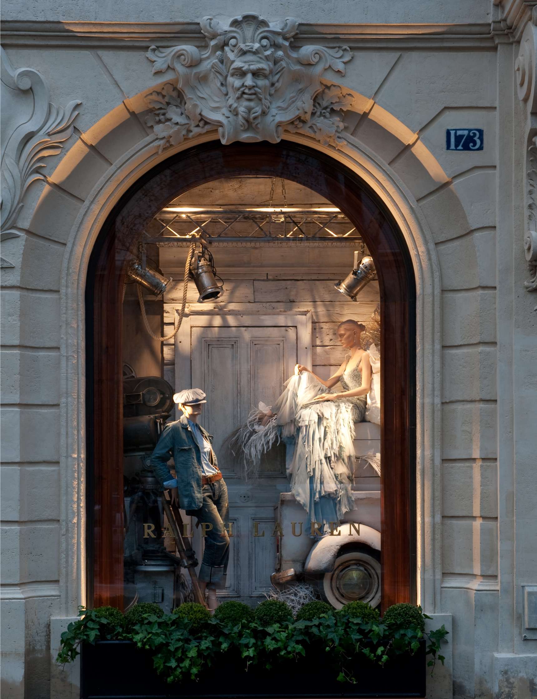 A decorative stone arch frames a store window display featuring mannequins in rustic, vintage-style clothing, with a background of wooden paneling and a potted plant below.