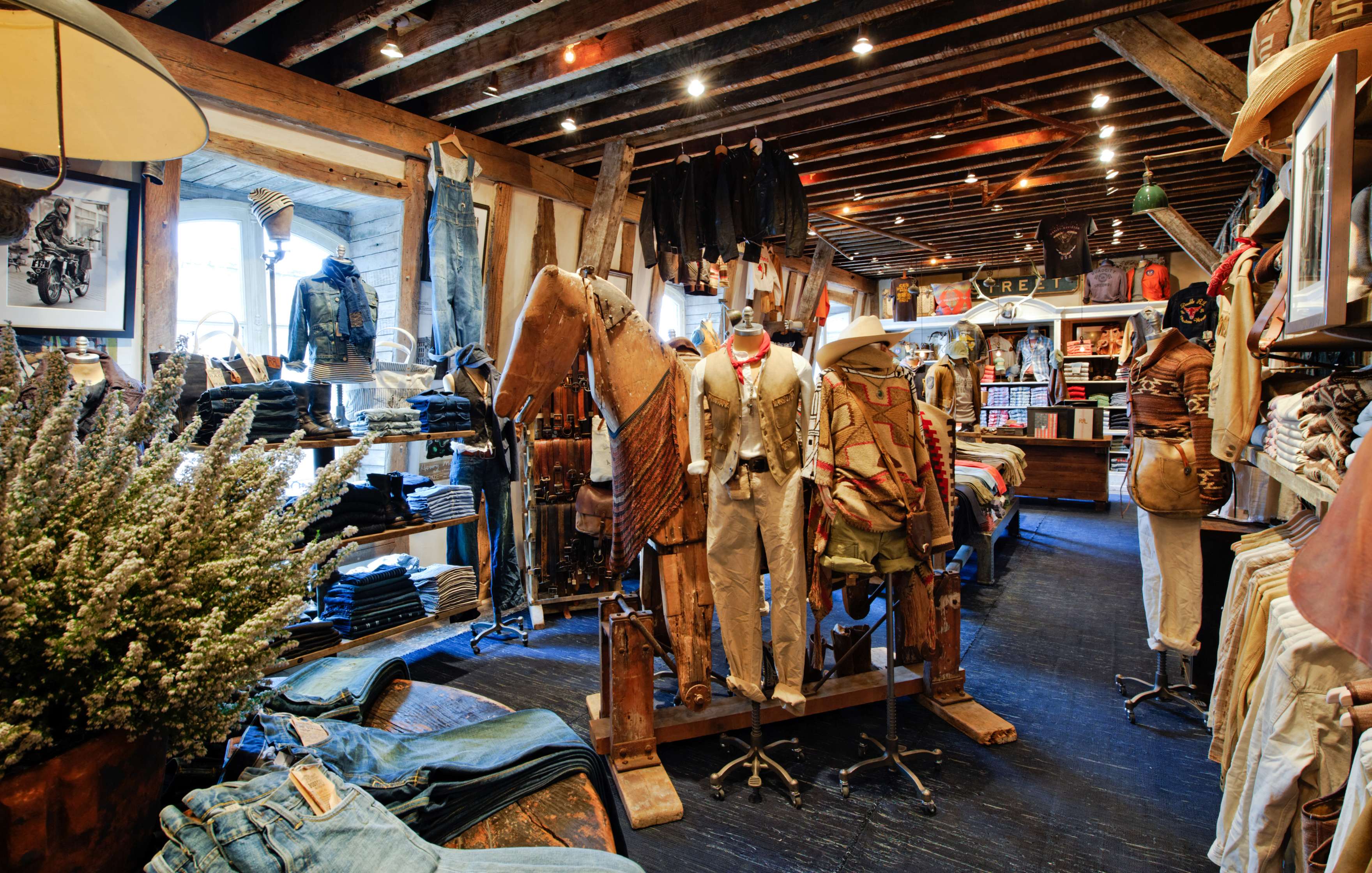 Interior of a clothing store with rustic decor, featuring various garments and accessories displayed on shelves, mannequins, and racks. A plant is positioned on the left side of the image.