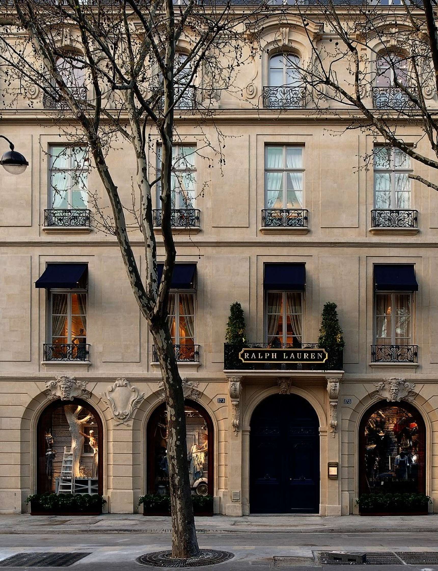 Street view of a Ralph Lauren store in a beige stone building with black awnings and large windows. Trees with bare branches stand in front.