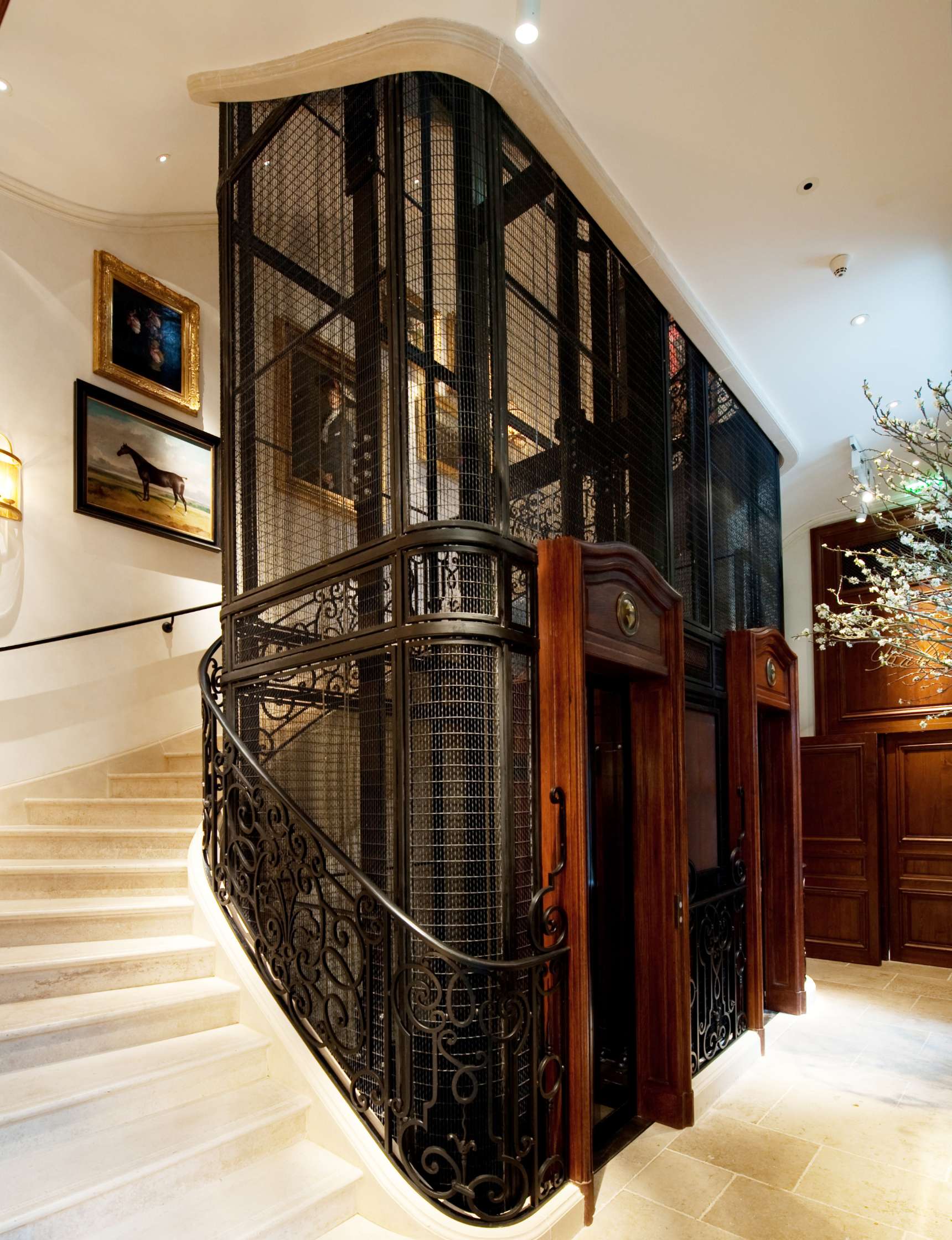 An ornate, vintage-style elevator with wrought iron and wood details is situated next to a staircase in an elegantly decorated interior space, featuring framed artwork on the walls.