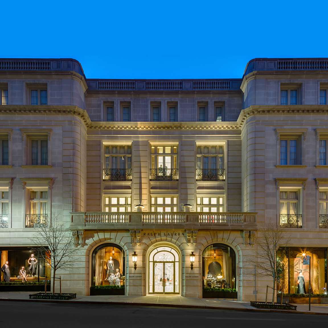 The image shows the facade of a grand, multi-story building with tall windows and ornate architectural details, illuminated at night. Mannequins are displayed in the ground floor windows.