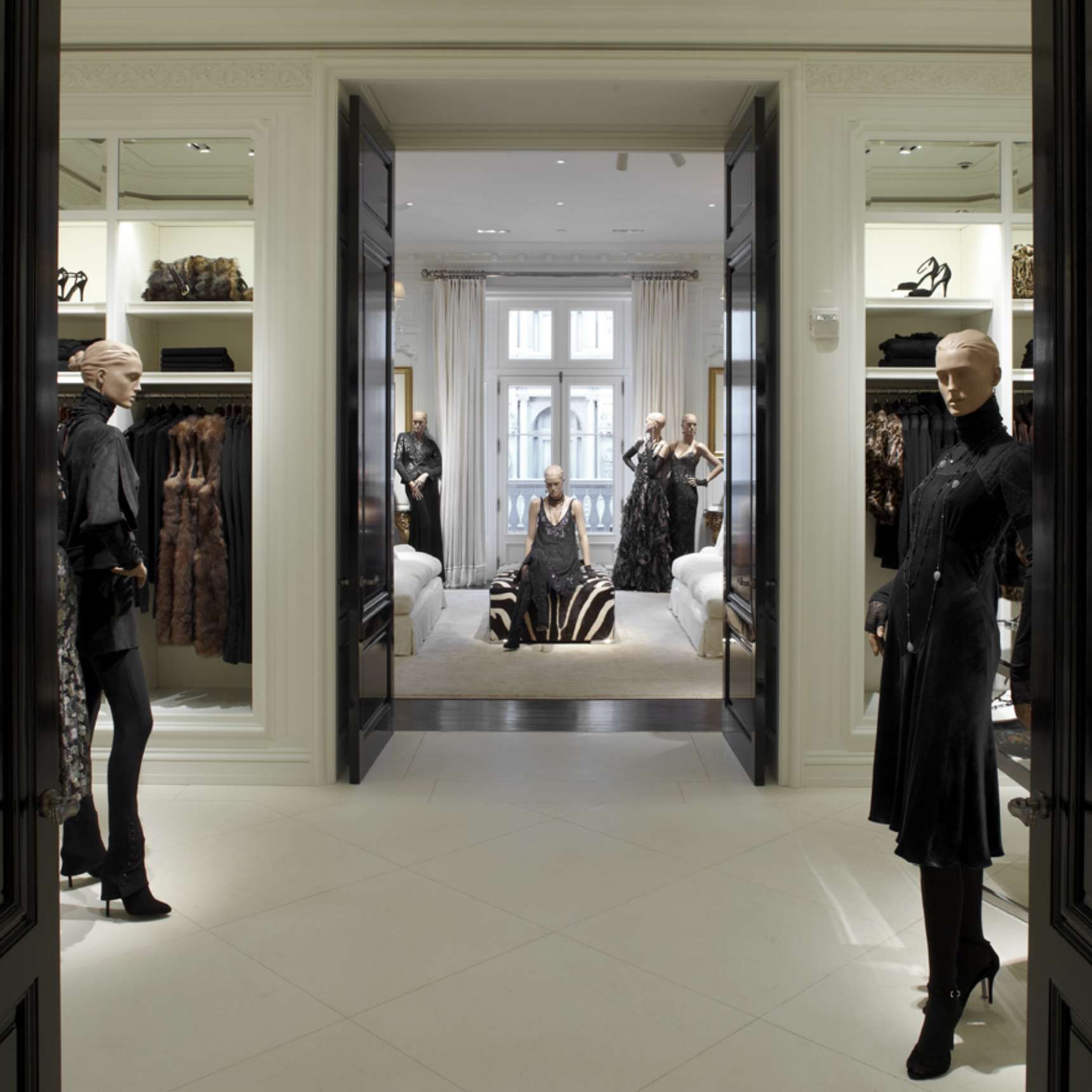 A high-end clothing store interior featuring mannequins dressed in dark outfits, with shelves of shoes and garments on either side and a bright, spacious area with more mannequins in the background.