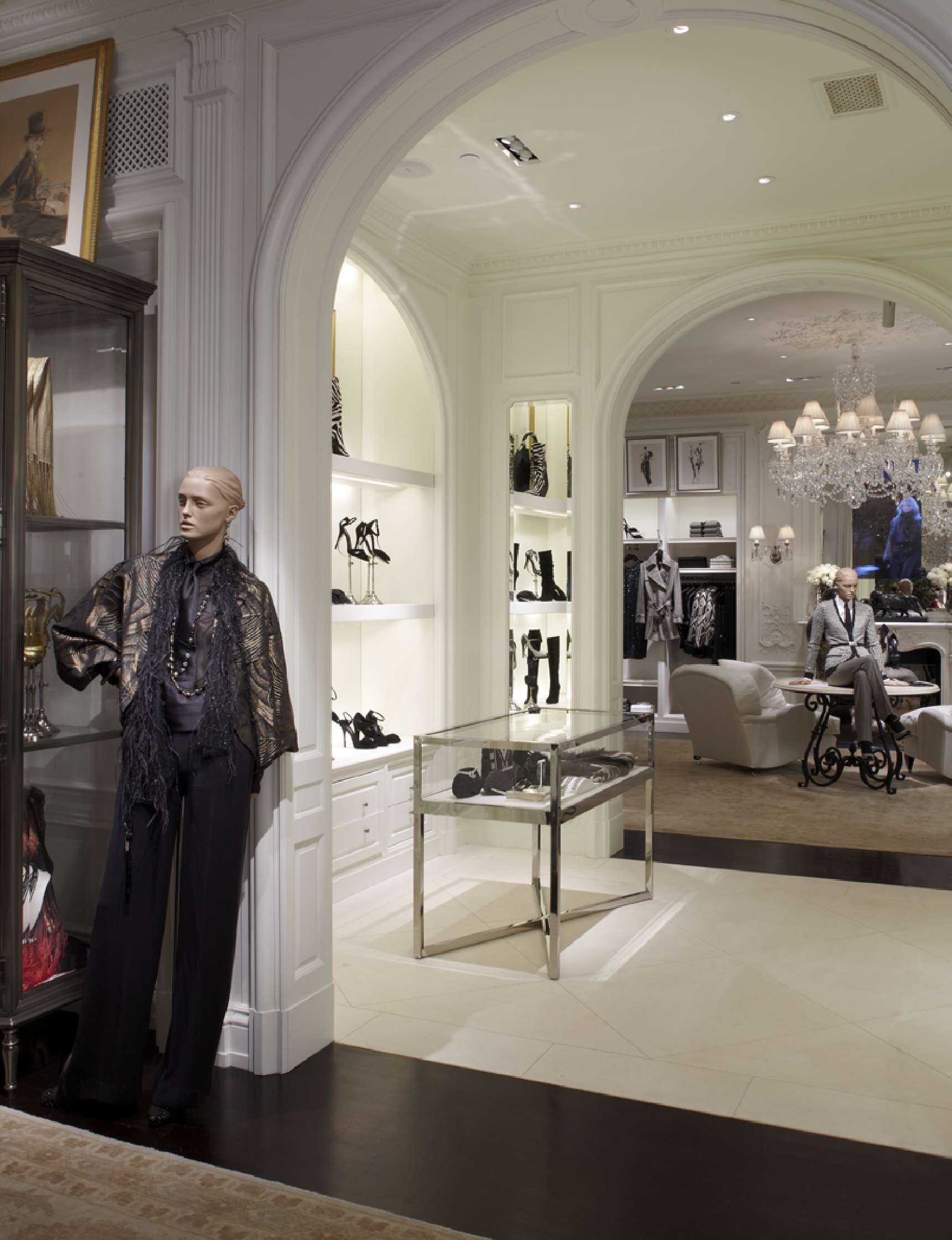 A luxury boutique interior with mannequins displaying clothing, shelves with shoes and accessories, glass display cases, and elegant chandeliers.