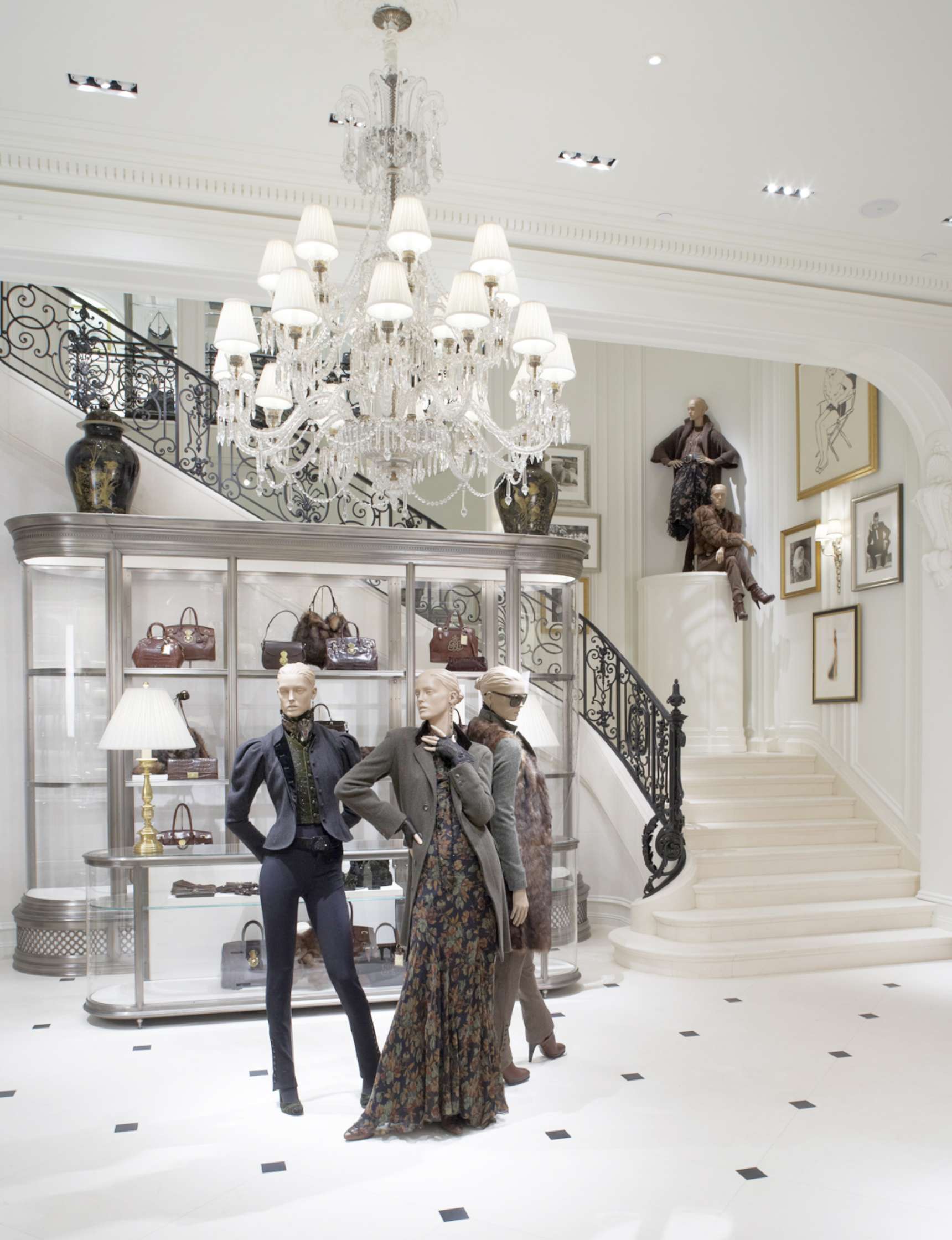 Luxurious retail store interior featuring mannequins in fashionable clothing, a grand staircase, a chandelier, handbags on display, and framed artwork on the walls.