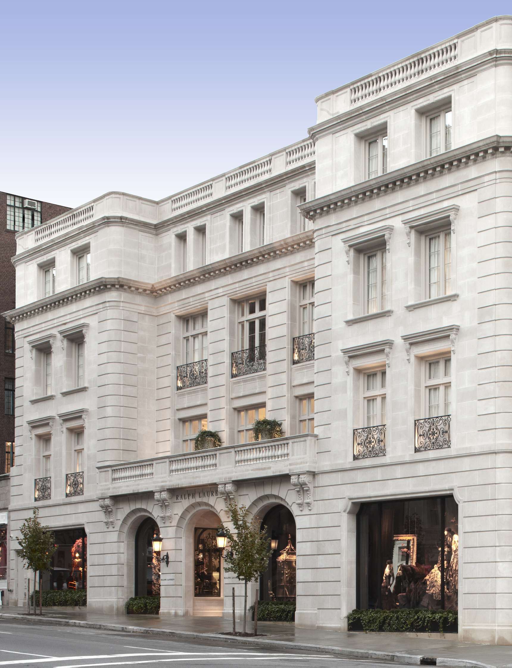 Exterior view of a multi-story stone building with large windows, ornate iron balconies, and entrance displaying a stylish retail store. Tree-lined sidewalk in front.