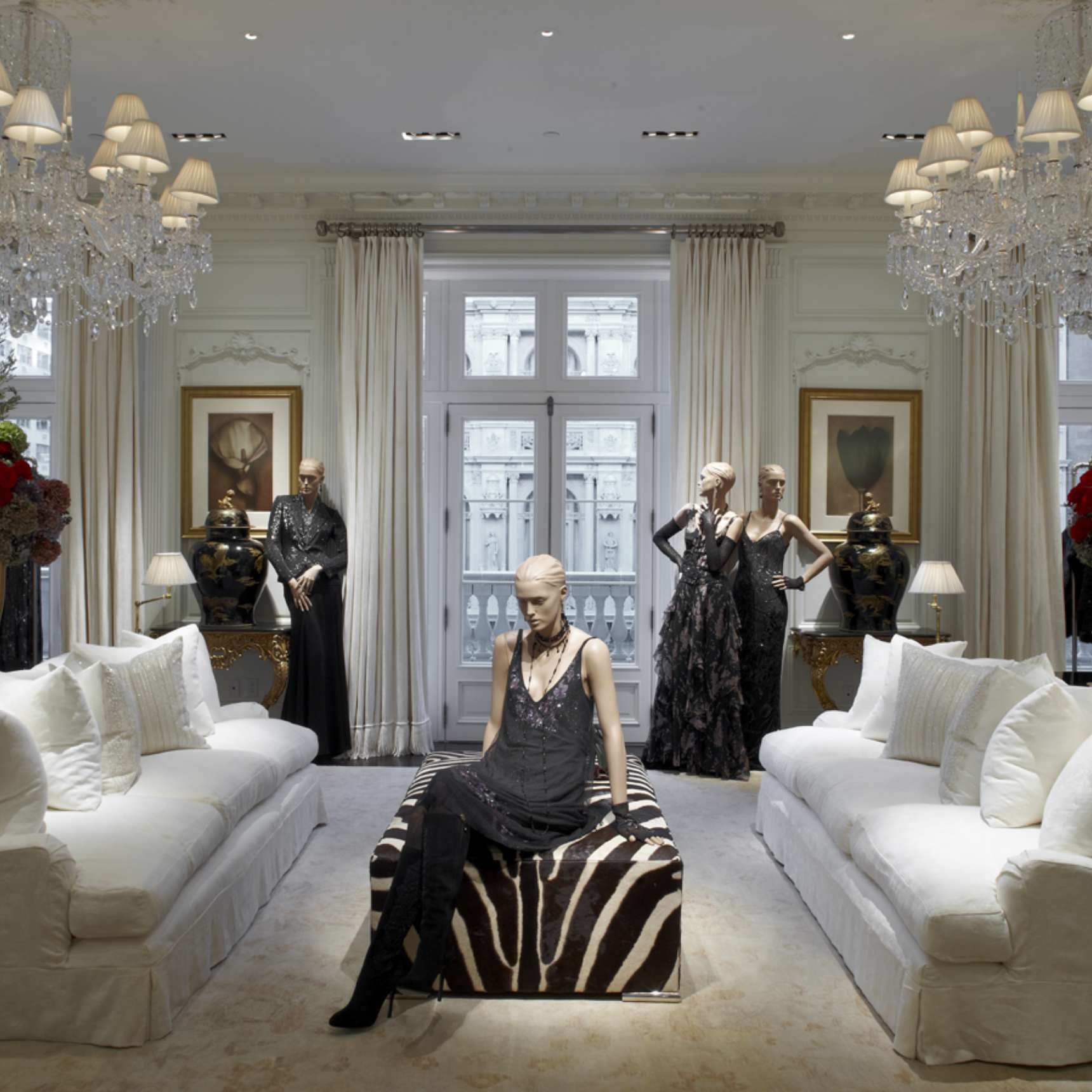 Three mannequins in elegant black dresses are displayed in a luxurious room with white sofas, chandeliers, and ornate decor.