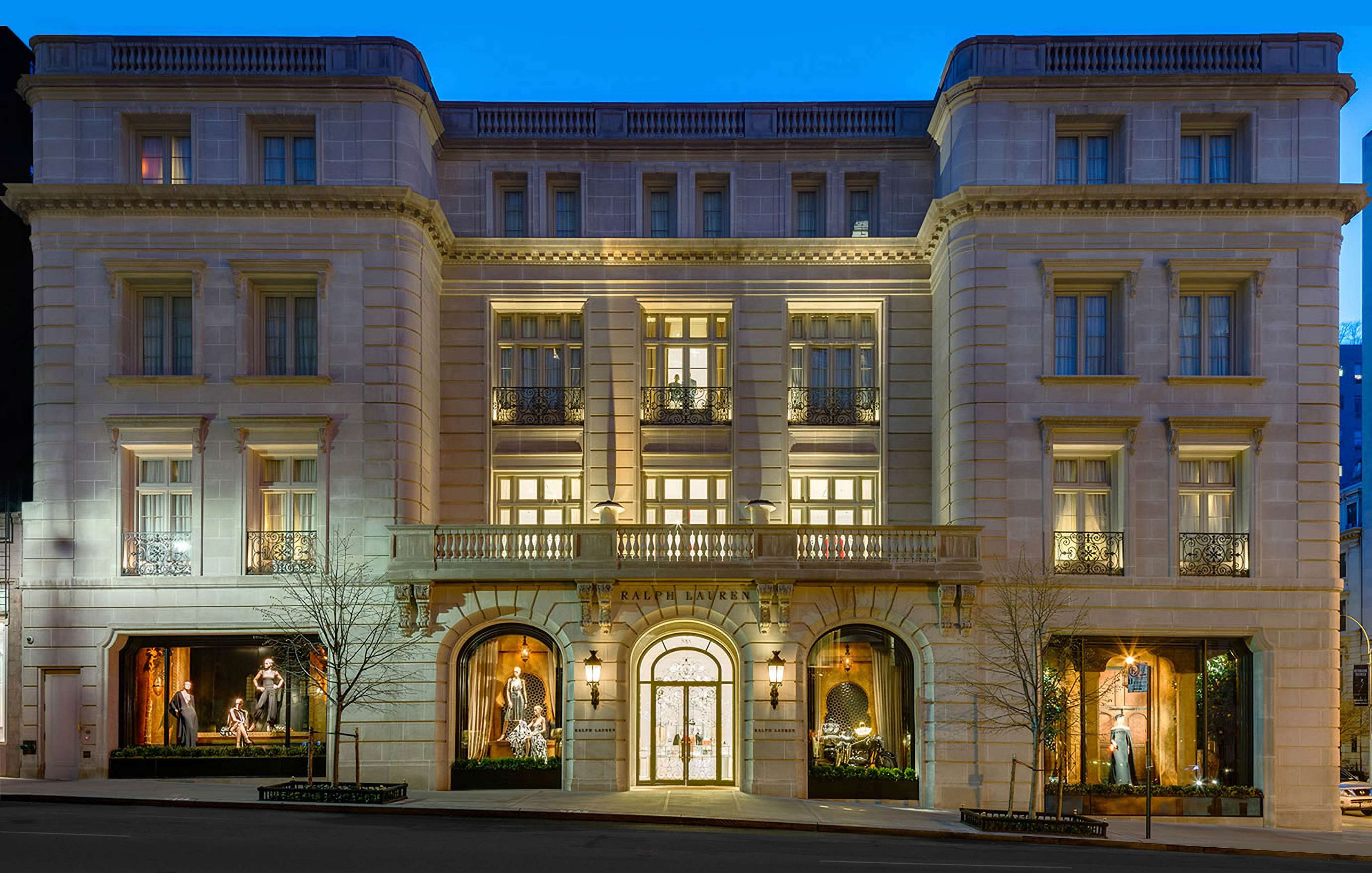 Elegant, well-lit three-story building facade with “Ralph Lauren” signage above main entrance. Large display windows showcase fashion items, and trees are sparsely planted in front. Evening scene.