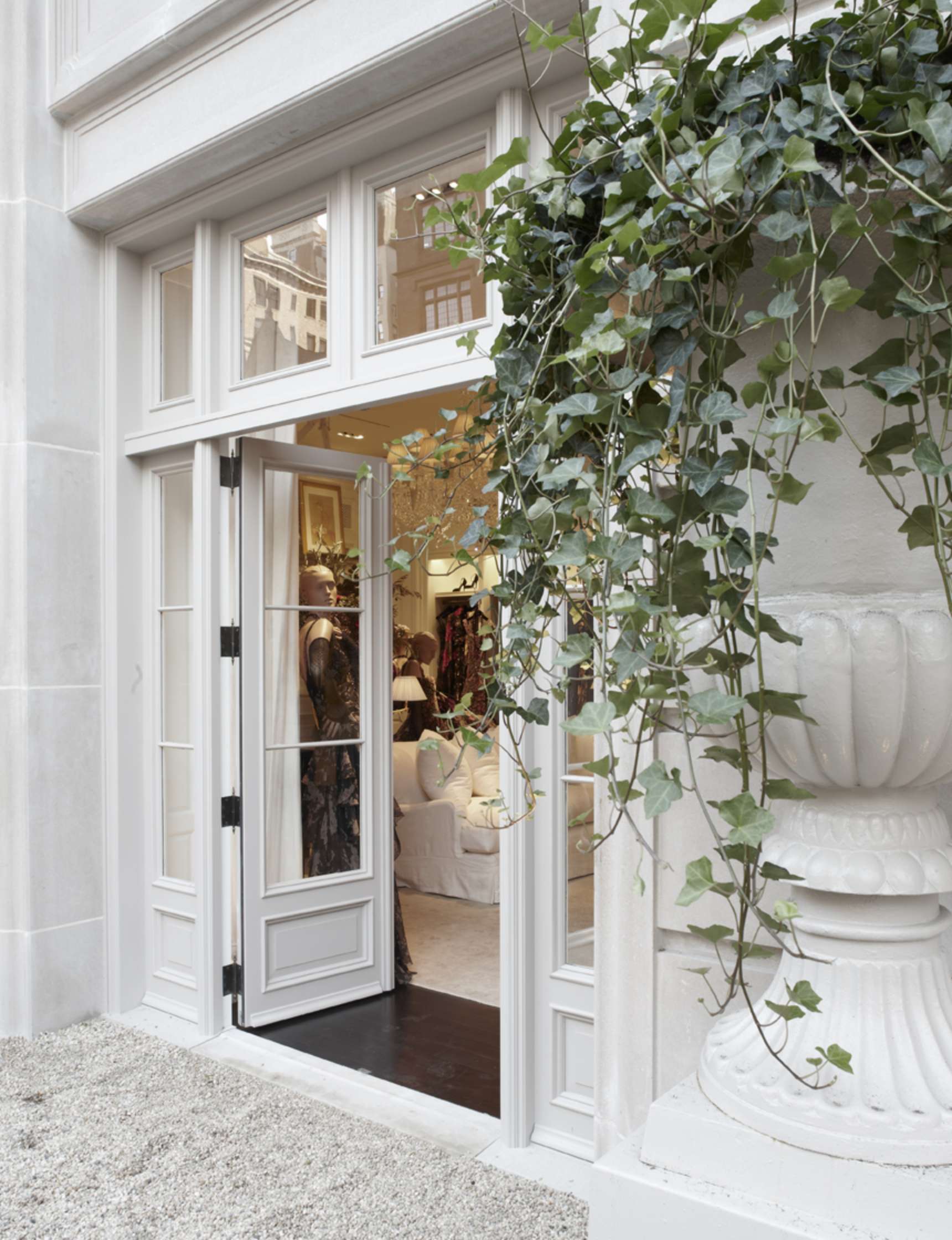 White double glass doors open to a brightly lit interior with mannequins displaying clothes. Ivy hangs from a large planter near the entrance.