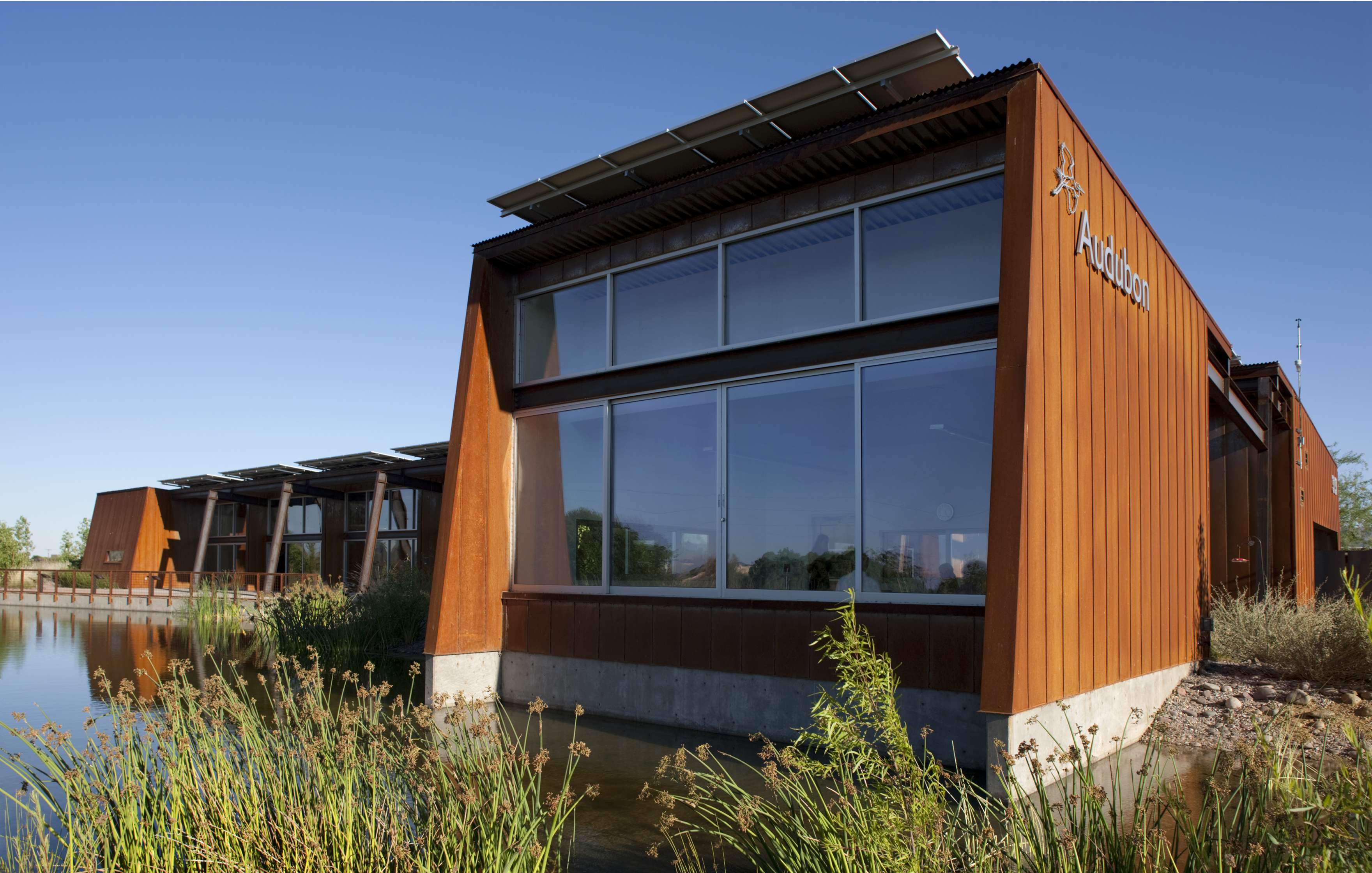 A modern building with large windows and rust-colored exterior stands next to a pond with reeds. Solar panels are visible on the roof. Another similar structure is seen in the background.