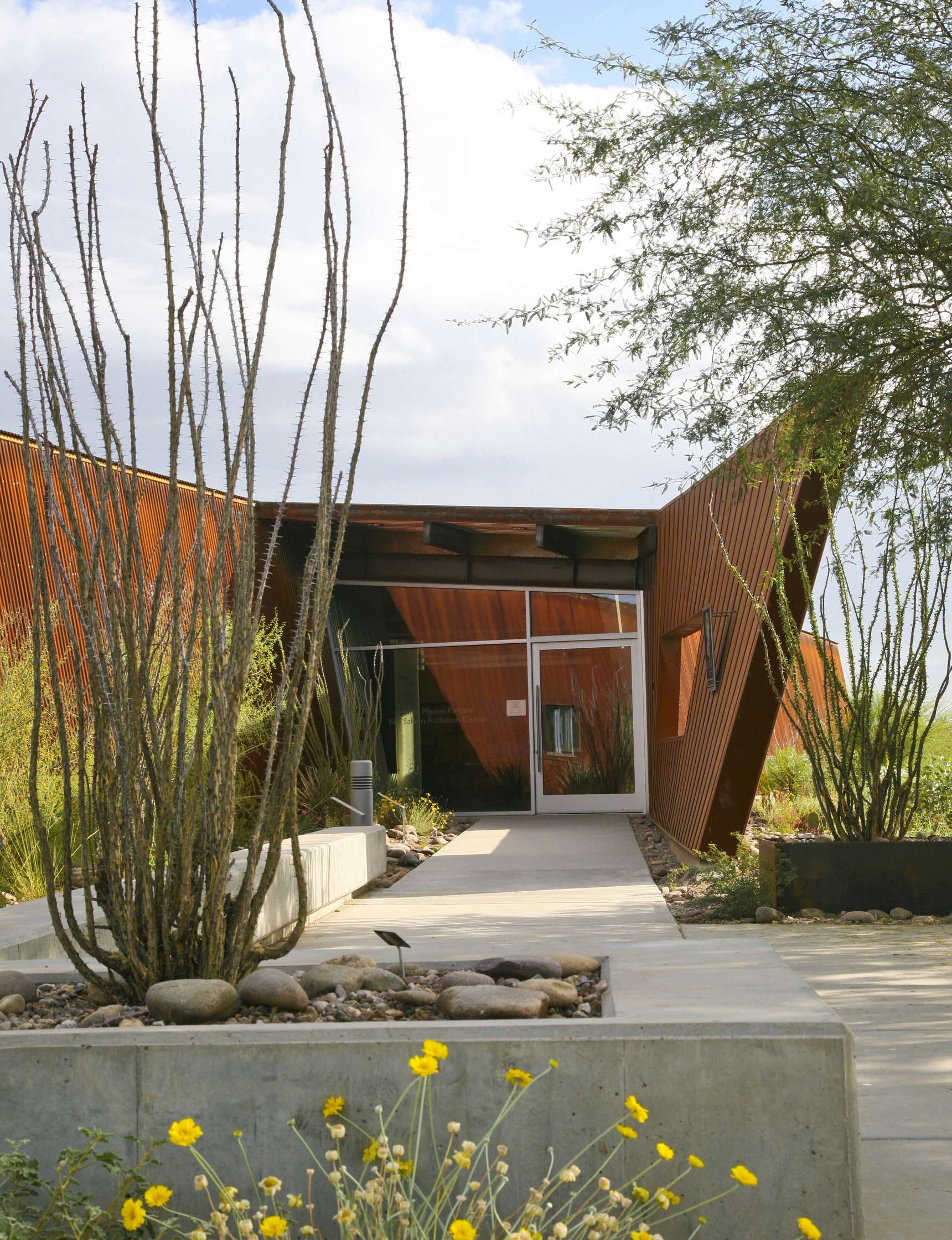 Modern building entrance with rust-colored metal exterior, large windows, and minimalist landscaping featuring tall, thin cacti and yellow flowers. Concrete walkway leads to the door.