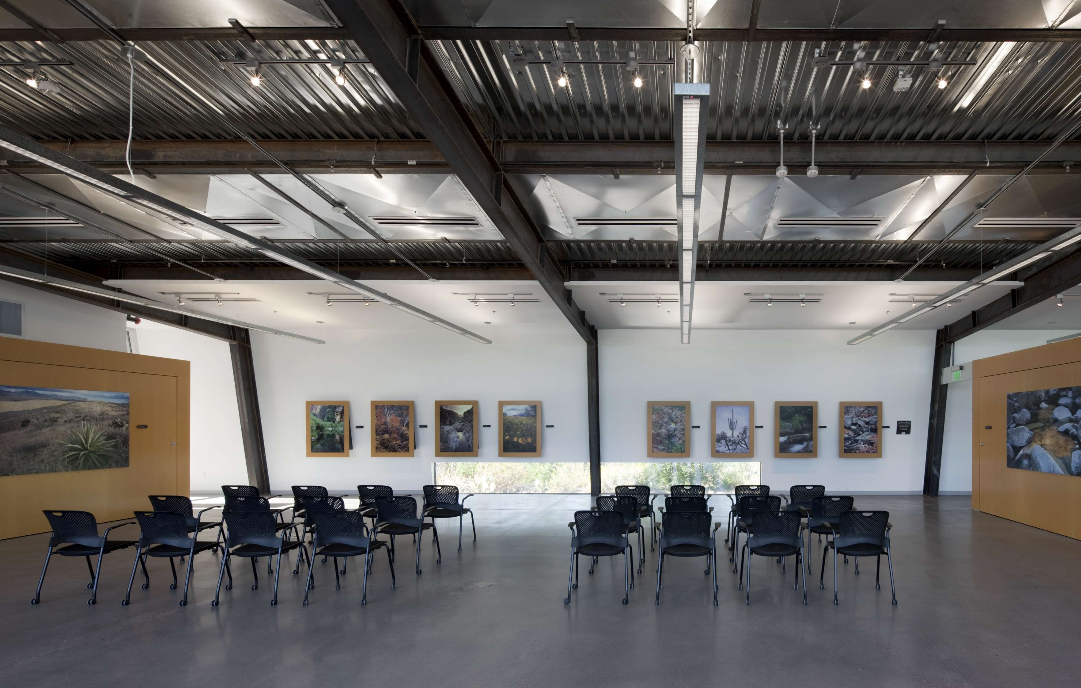 Modern gallery with industrial ceiling design, featuring wall-mounted photographs and rows of black chairs facing the display.