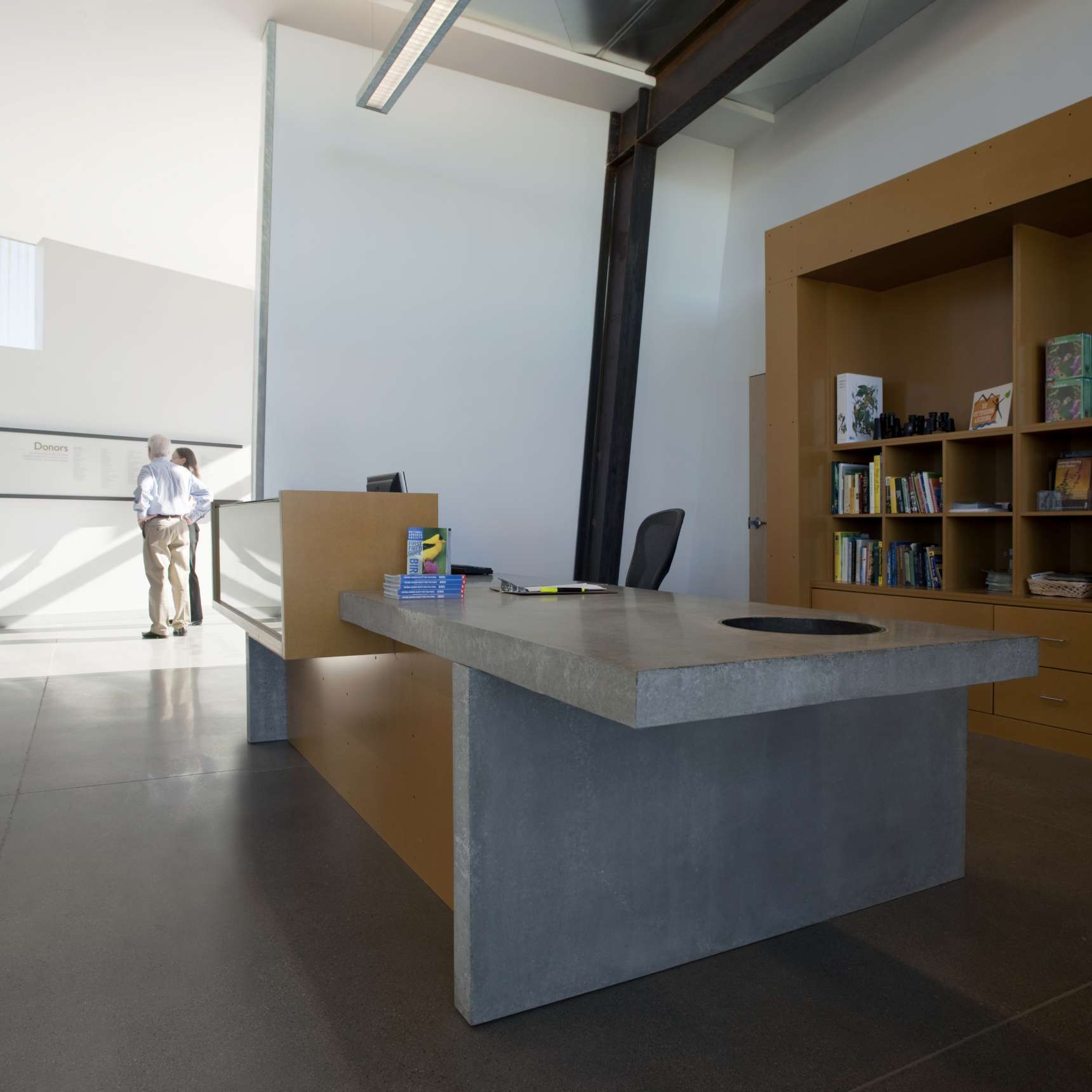 A modern office with a concrete desk in the foreground, a bookshelf with books and toys on the right, and a person standing near a wall display in the background.