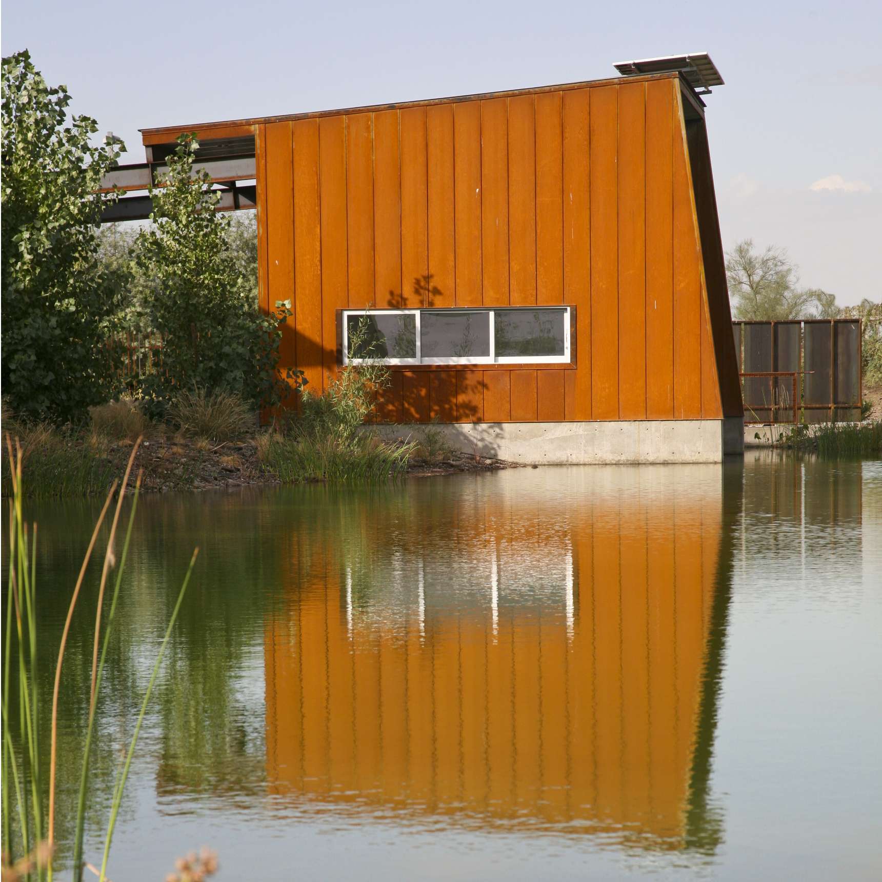 A small rust-colored building with a slanted roof sits next to a still body of water, reflecting the structure. There's a rectangular window and greenery around the building.