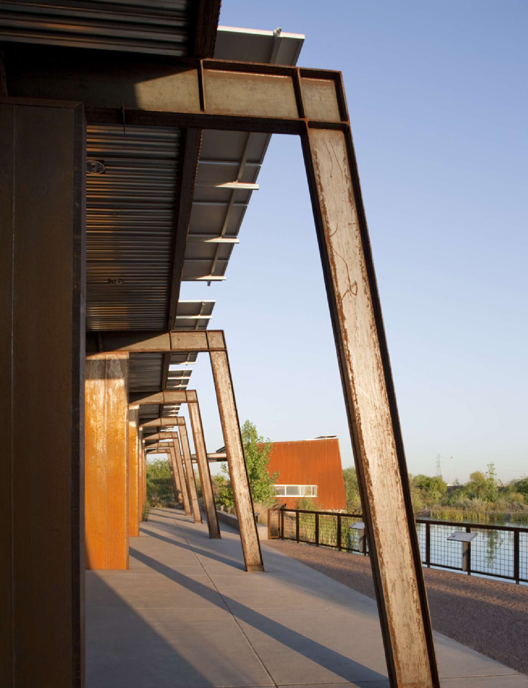 A walkway with rust-colored metal beams and a corrugated metal roof, adjacent to a body of water, under a clear sky.