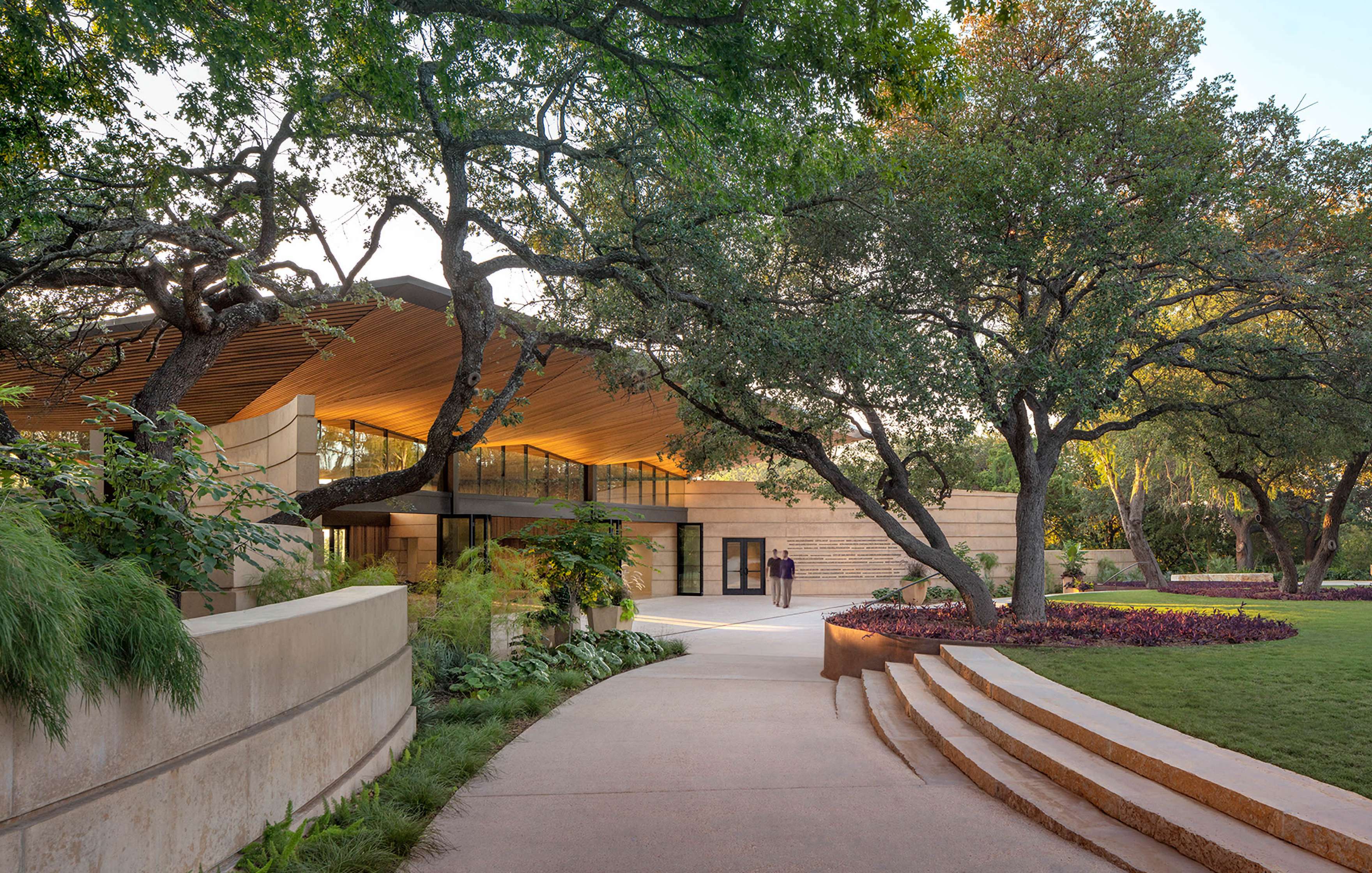 Modern architectural building with large overhang, surrounded by trees and well-maintained landscaped garden, with two individuals standing near the entrance.