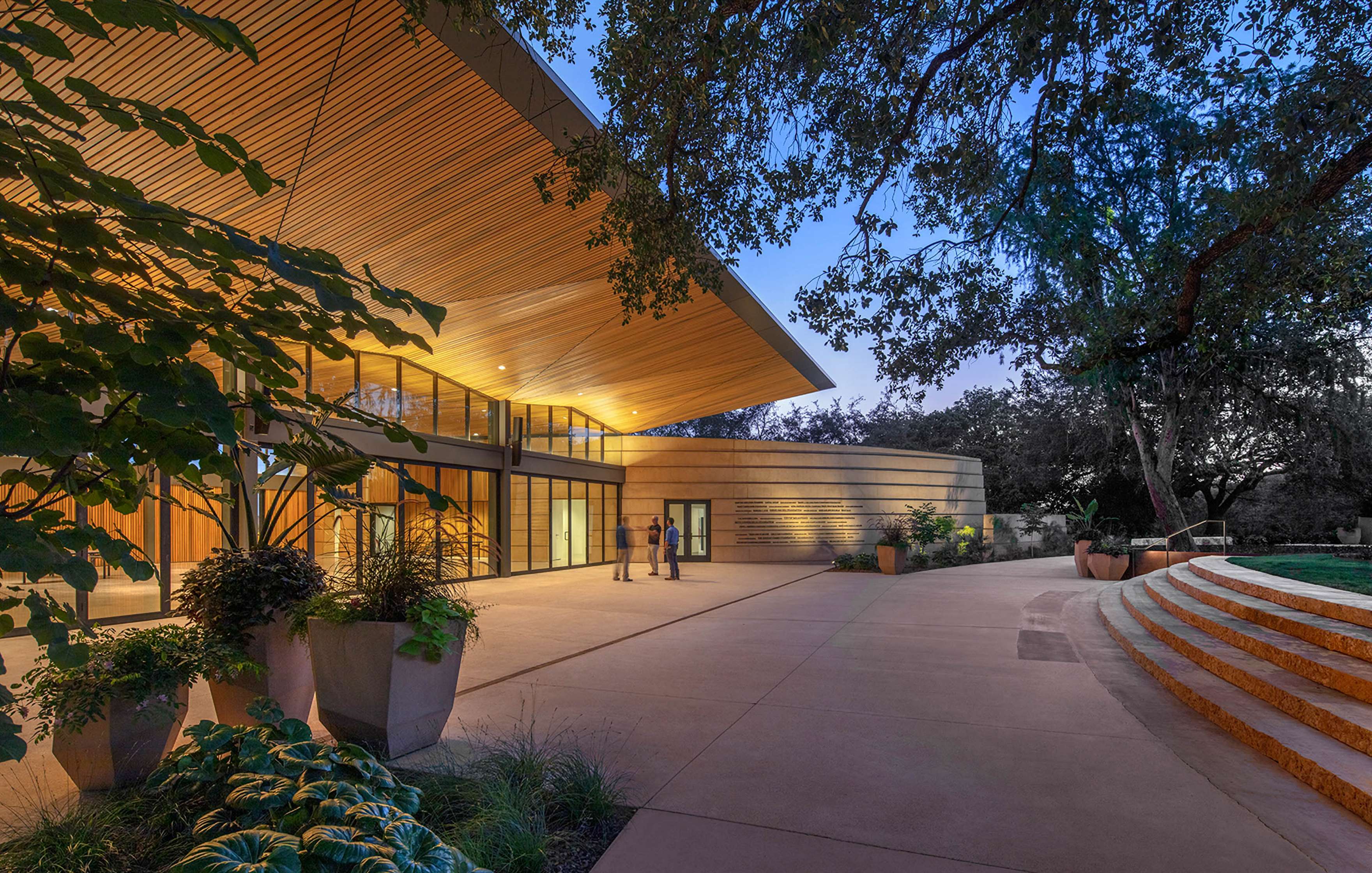 A modern building with a wooden overhang and large glass windows, surrounded by plants and trees. The courtyard features steps leading up to the entrance. Two people stand near the entrance.