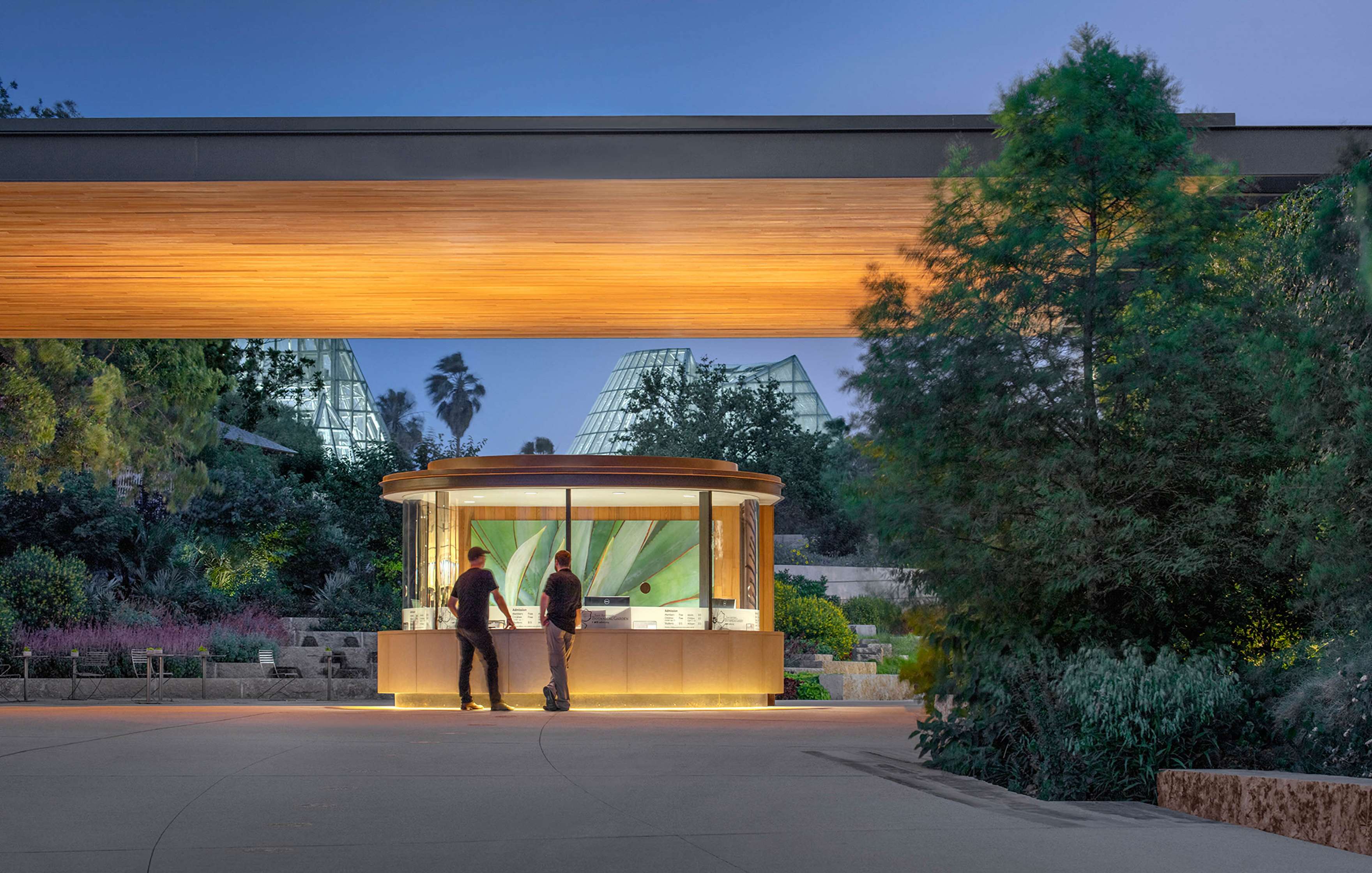 Two people standing in front of a small, illuminated kiosk under a wooden overhang in a botanical garden setting with trees and glass pyramid structures in the background.