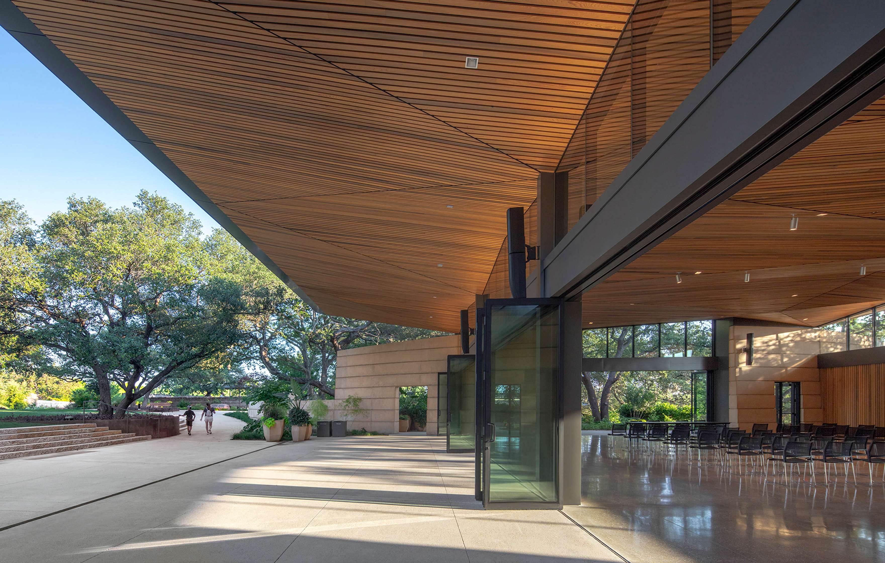 A modern building with a large wooden overhang and glass doors overlooks a courtyard with trees and greenery.