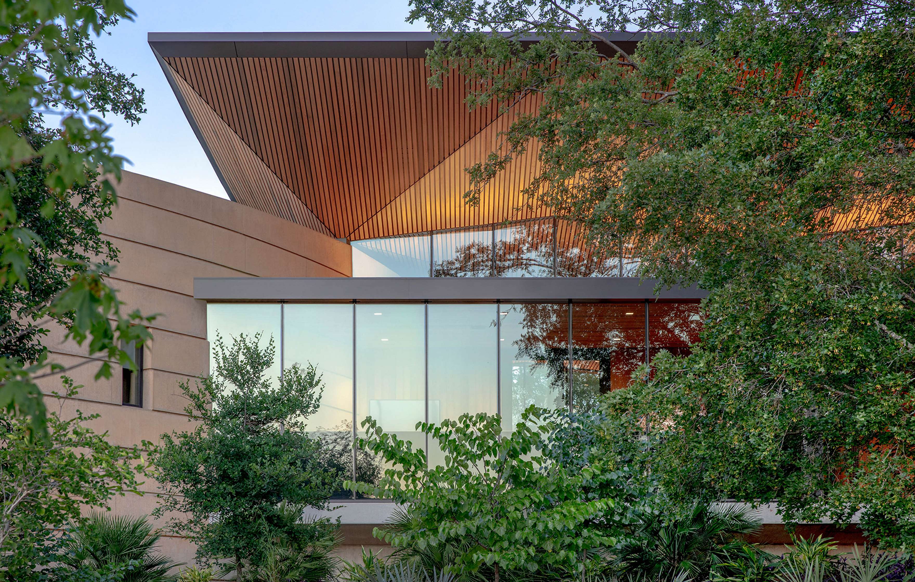 A modern building with large glass windows is partially obscured by lush green trees. The roof has an upward slant and wooden paneling visible underneath.